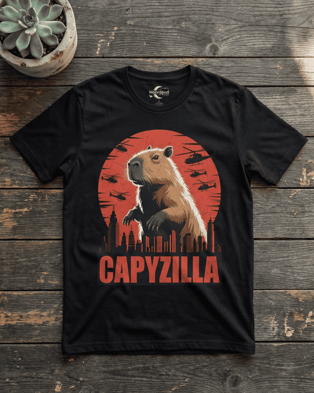 A black Funny Capybara Monster T-Shirt laid flat on a rustic wooden surface, featuring a bold 'CAPYZILLA' Godzilla Parody graphic. The design depicts a colossal capybara standing over a city skyline against a dramatic red sunset, with several small helicopters flying in the background. A green succulent plant sits in a concrete pot in the upper left, adding to the visual appeal of this engaging graphic tee.