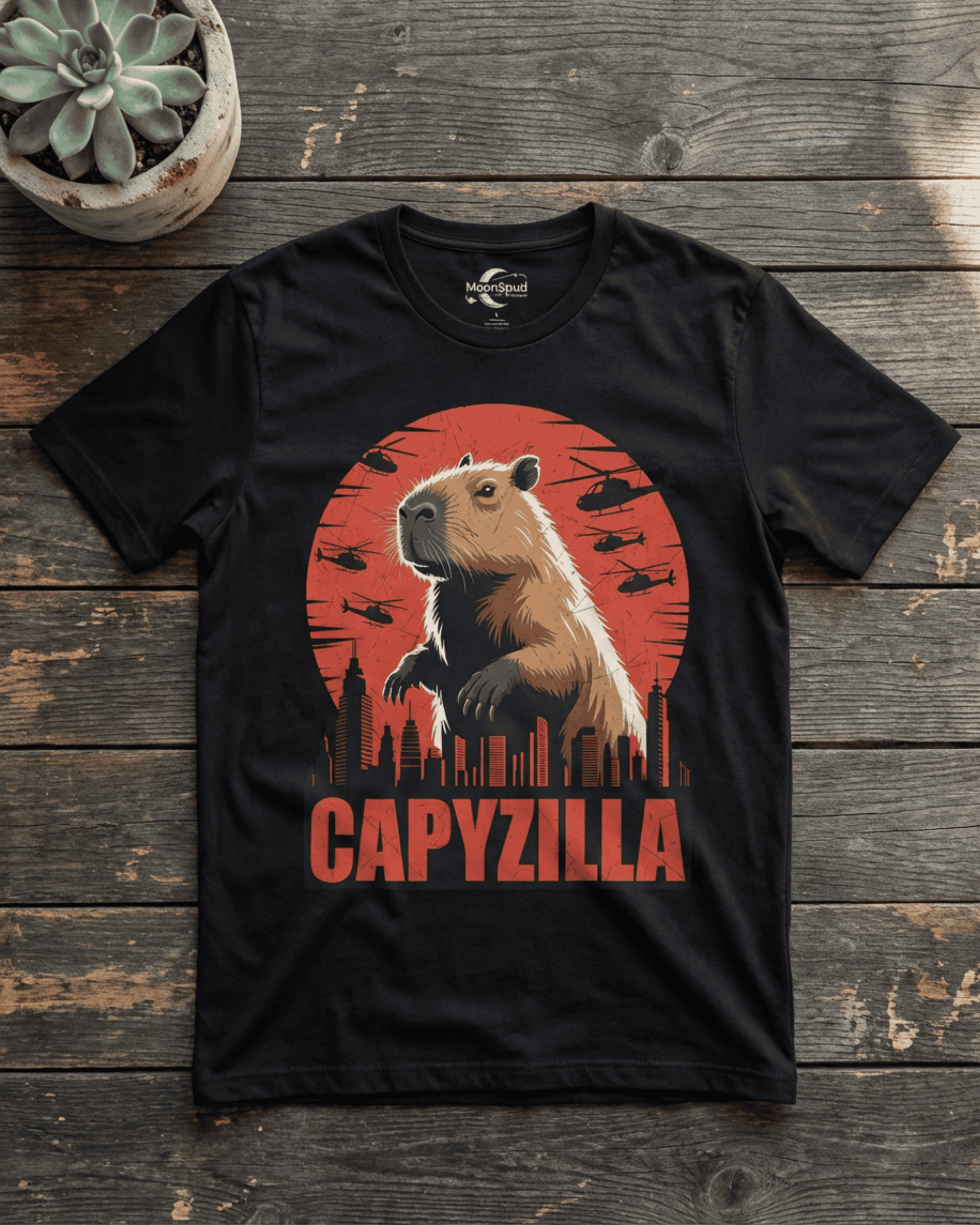 A black Funny Capybara Monster T-Shirt laid flat on a rustic wooden surface, featuring a bold 'CAPYZILLA' Godzilla Parody graphic. The design depicts a colossal capybara standing over a city skyline against a dramatic red sunset, with several small helicopters flying in the background. A green succulent plant sits in a concrete pot in the upper left, adding to the visual appeal of this engaging graphic tee.