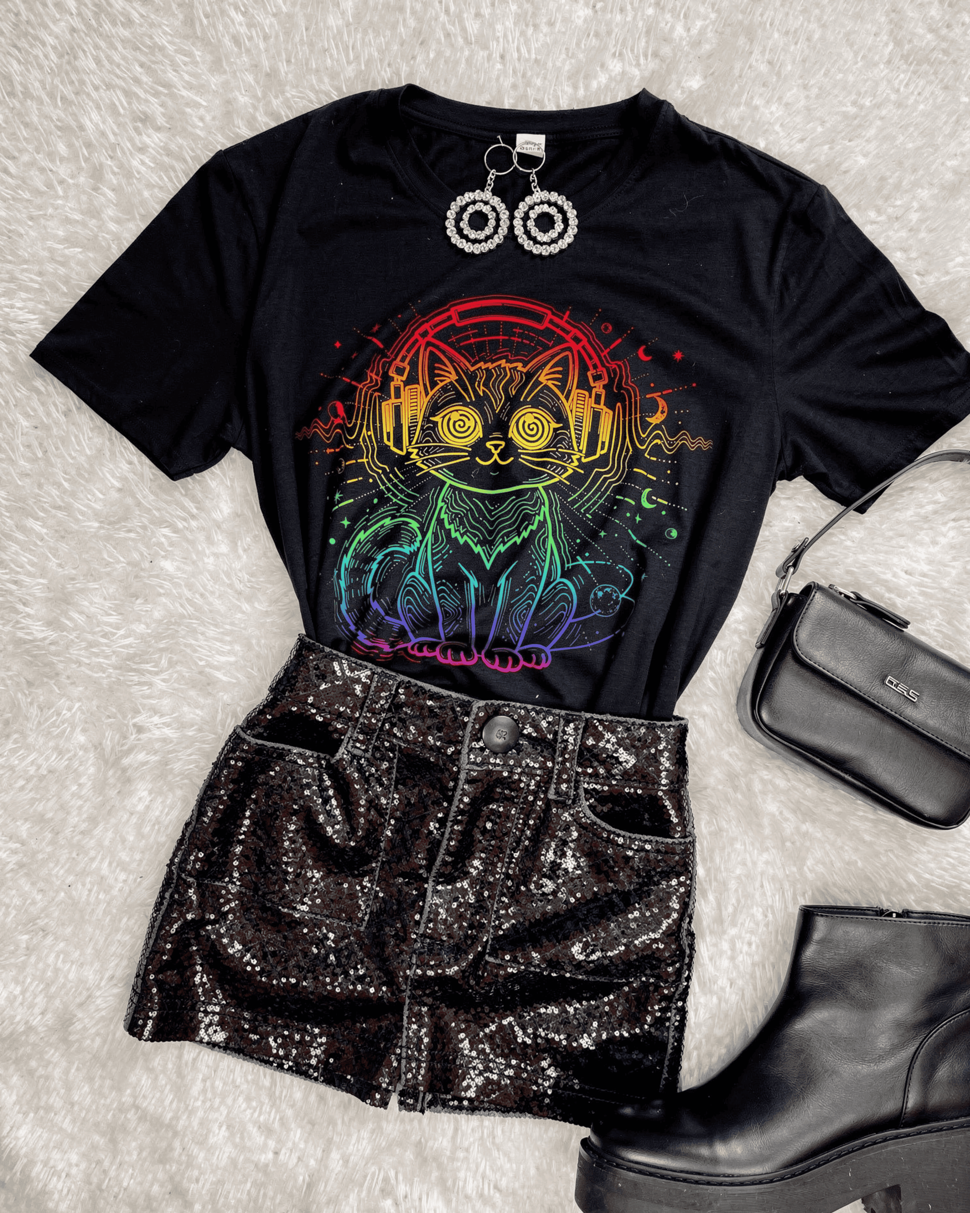 A fashion flat lay of a complete festival outfit centered around a black Music Lover DJ Cat T-Shirt. The shirt's vibrant, retro rave EDM graphic is paired with a black sequin skirt, platform boots, and a purse on a white shag rug.