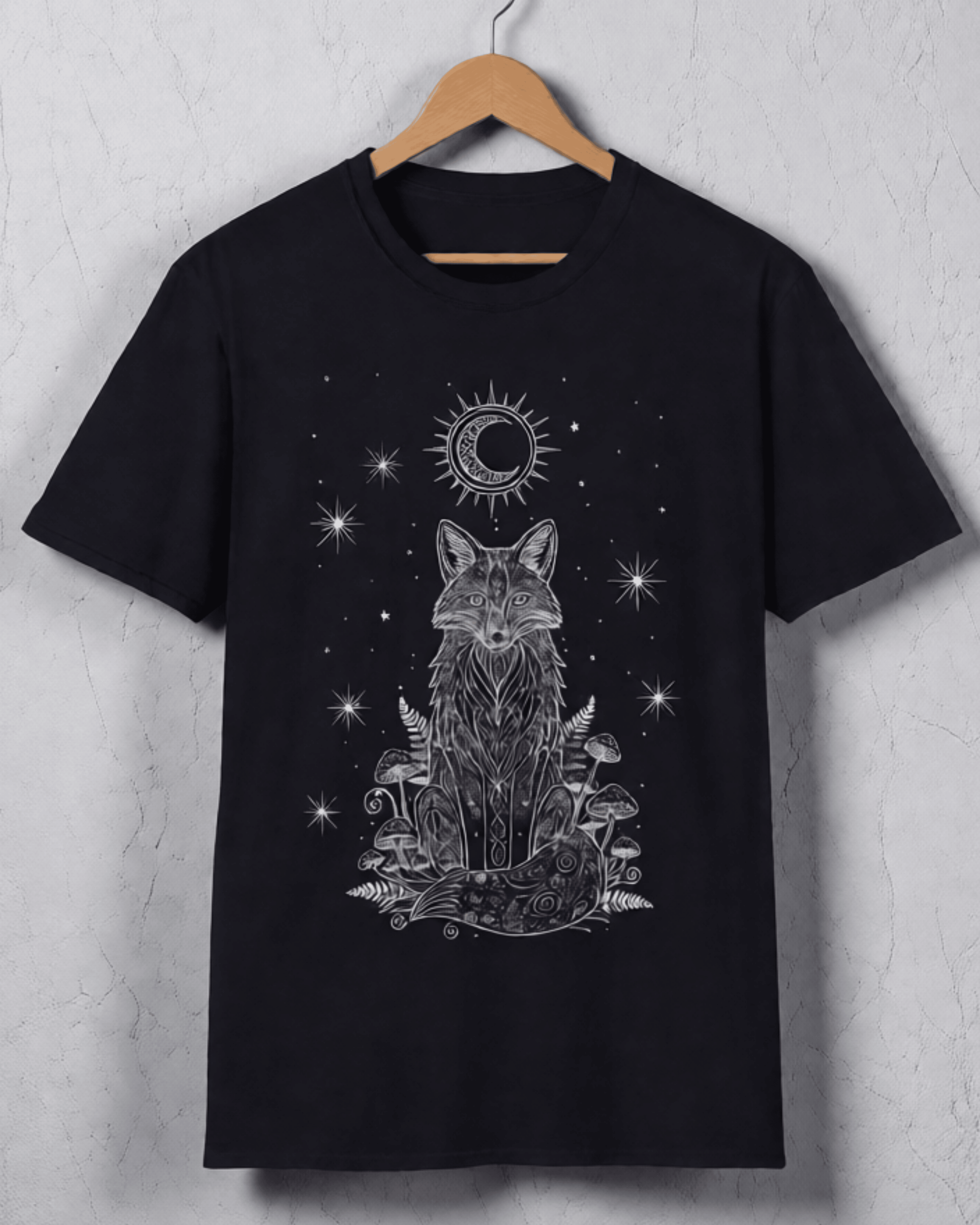 A Black T-Shirt hanging against a textured light grey wall, clearly displaying the front graphic. The Celestial Tshirt design highlights a geometric fox surrounded by ferns and mushrooms under a moon phase, serving as a versatile Witchy Crescent Moon Graphic Tee.
