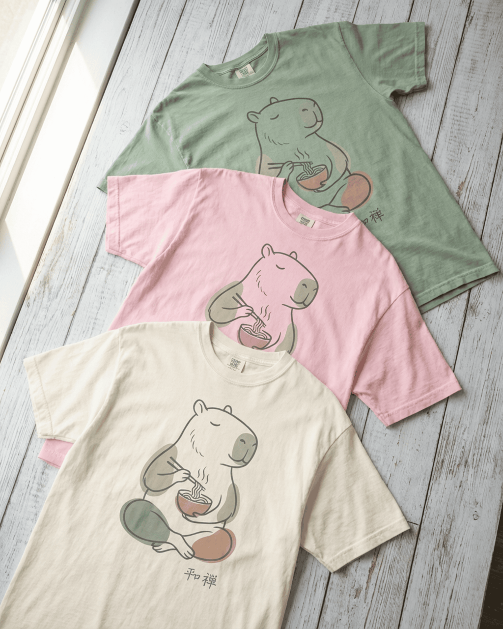 An overhead view of three folded Capybara Peaceful Zen Ramen T-Shirts arranged on a light wood background. This image highlights the soft, heavyweight cotton of the Comfort Colors 1717 tee and displays the full color palette of Ivory, Blossom pink, and Bay green for this serene design.