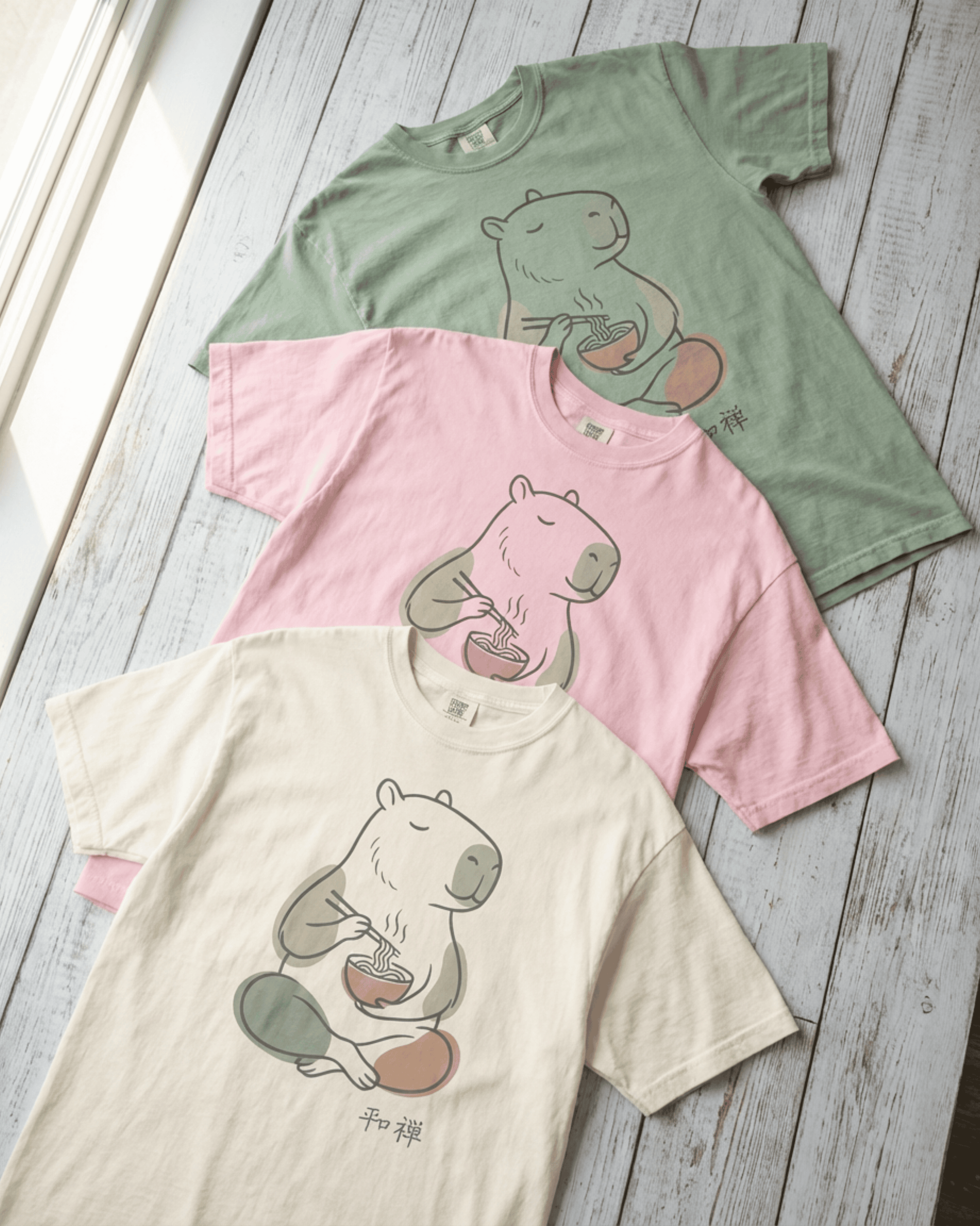 An overhead view of three folded Capybara Peaceful Zen Ramen T-Shirts arranged on a light wood background. This image highlights the soft, heavyweight cotton of the Comfort Colors 1717 tee and displays the full color palette of Ivory, Blossom pink, and Bay green for this serene design.