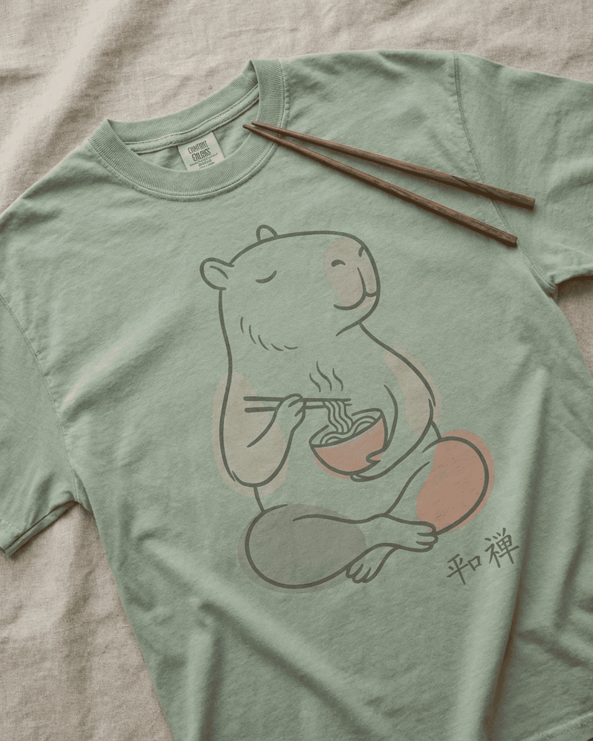 A flat lay of the Capybara Peaceful Zen Ramen T-Shirt in the color Bay. This soft, garment-dyed Comfort Colors 1717 tee features a charming line-art illustration of a capybara in a meditative pose, contentedly eating a steaming bowl of ramen with chopsticks. Below the tranquil design are the Japanese kanji for 'Peace and Zen'.