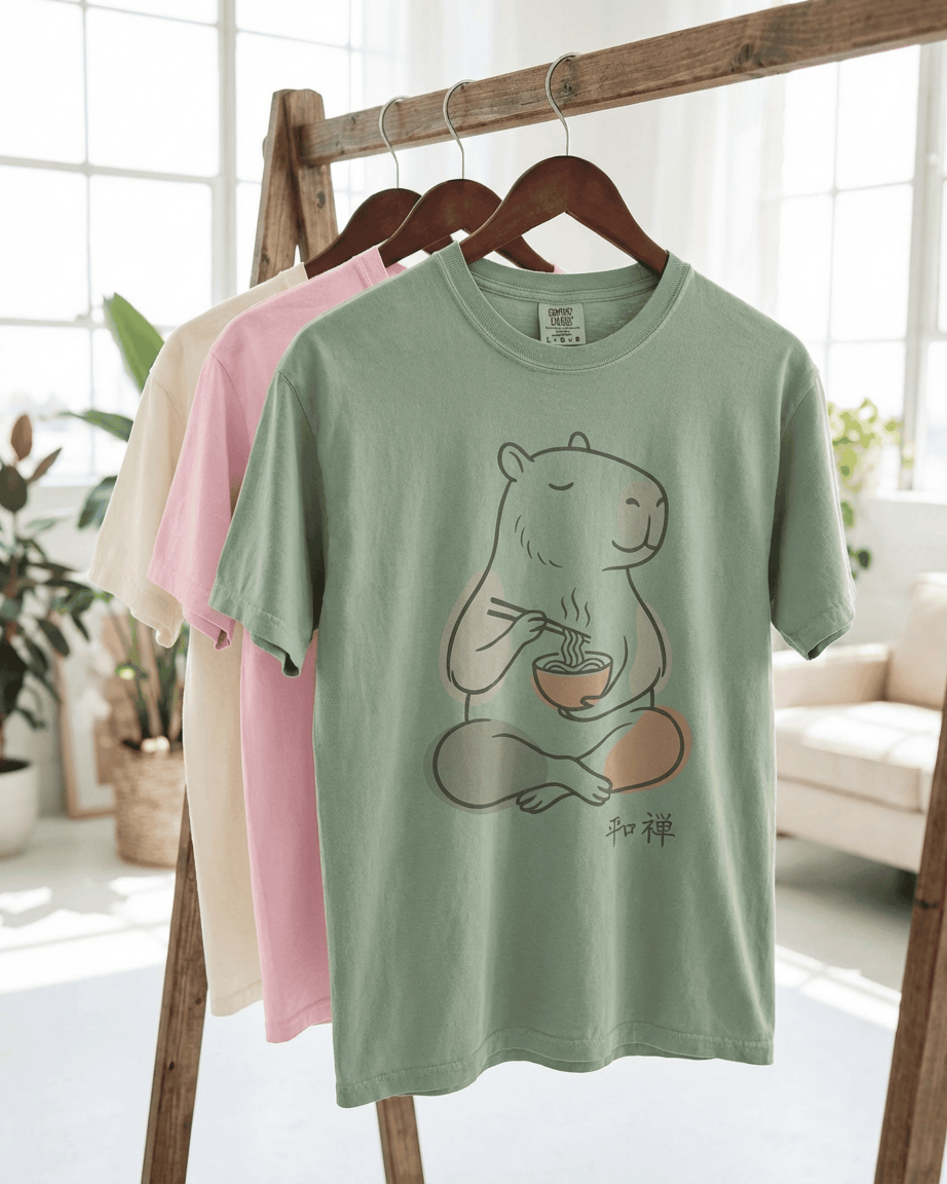 Showcasing the color options for the Capybara Peaceful Zen Ramen T-Shirt. Three premium Comfort Colors 1717 tees in Bay green, Blossom pink, and Ivory hang on a wooden clothing rack in a bright, airy studio, allowing customers to see the variety available for this unique design.