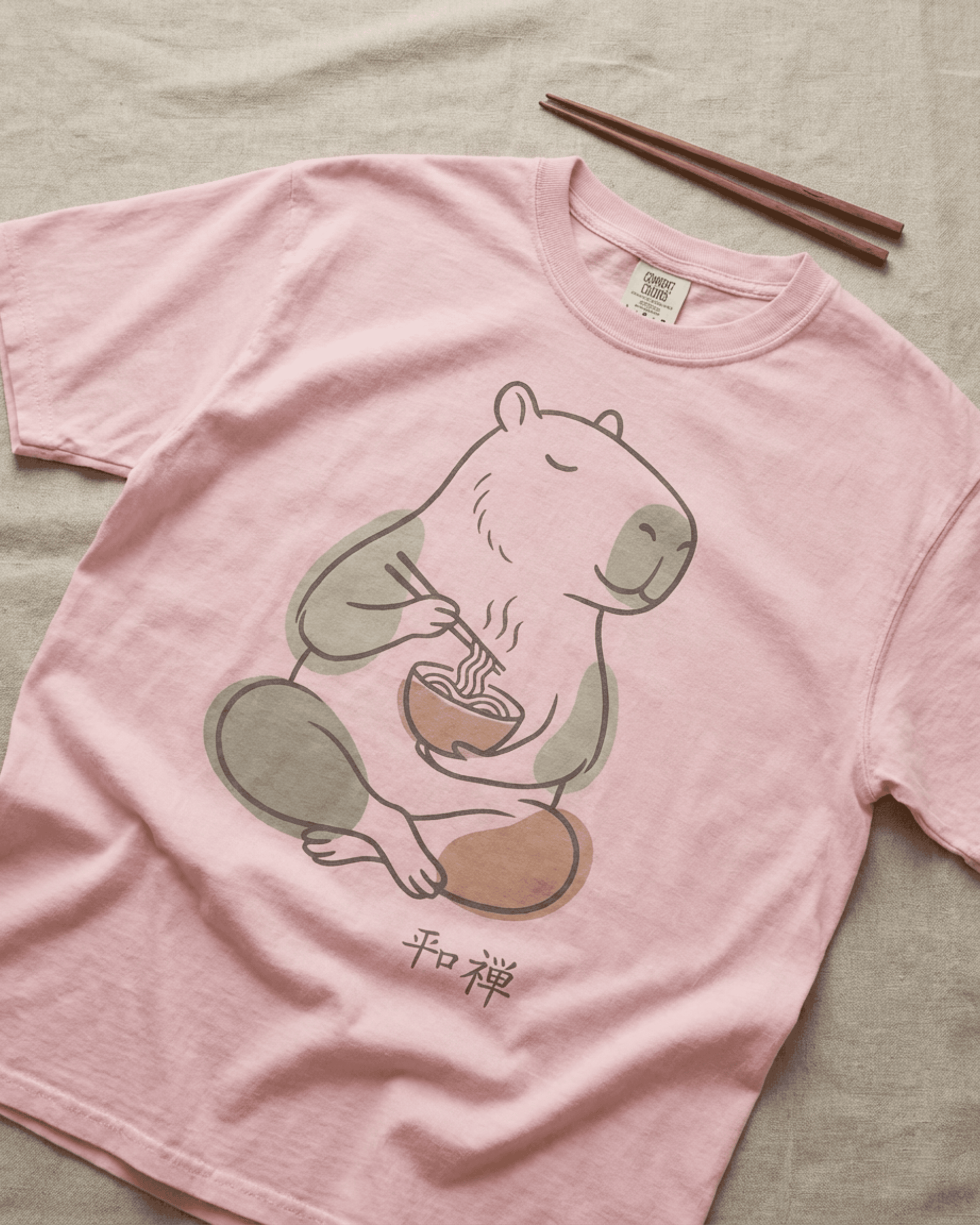 Flat lay of the Capybara Peaceful Zen Ramen T-Shirt in a beautiful Blossom pink. This relaxed-fit Comfort Colors 1717 tee is styled on a linen background with a pair of dark wood chopsticks beside the collar, perfectly complementing the ramen-eating capybara design.