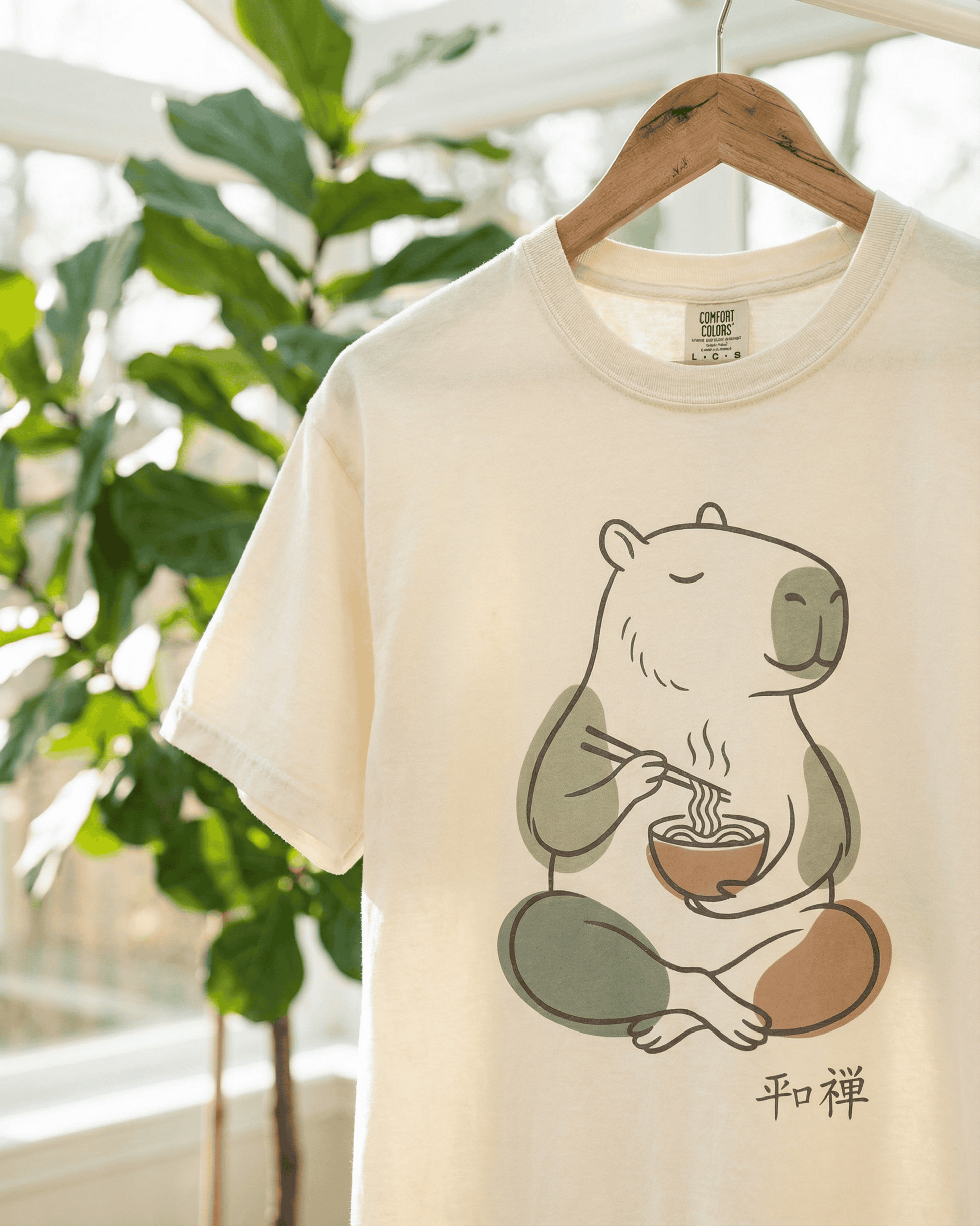 Close-up of the Ivory Capybara Peaceful Zen Ramen T-Shirt hanging on a wooden hanger. The soft, garment-dyed Comfort Colors 1717 tee features a minimalist design of a capybara meditating while eating a bowl of steaming ramen with chopsticks.