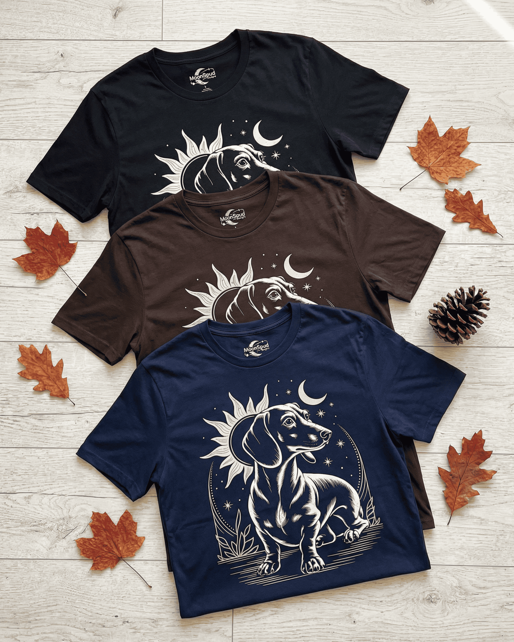 A flat lay of three celestial-themed Dachshund T-shirts in black, chocolate brown, and navy blue. Each beautiful weiner dog shirt features a screen-printed graphic of a long dog sitting proudly under a sun and crescent moon, making it a perfect gift for any dog lover, dog mom, or dog dad. The shirts are arranged on a light wood background with scattered autumn leaves.