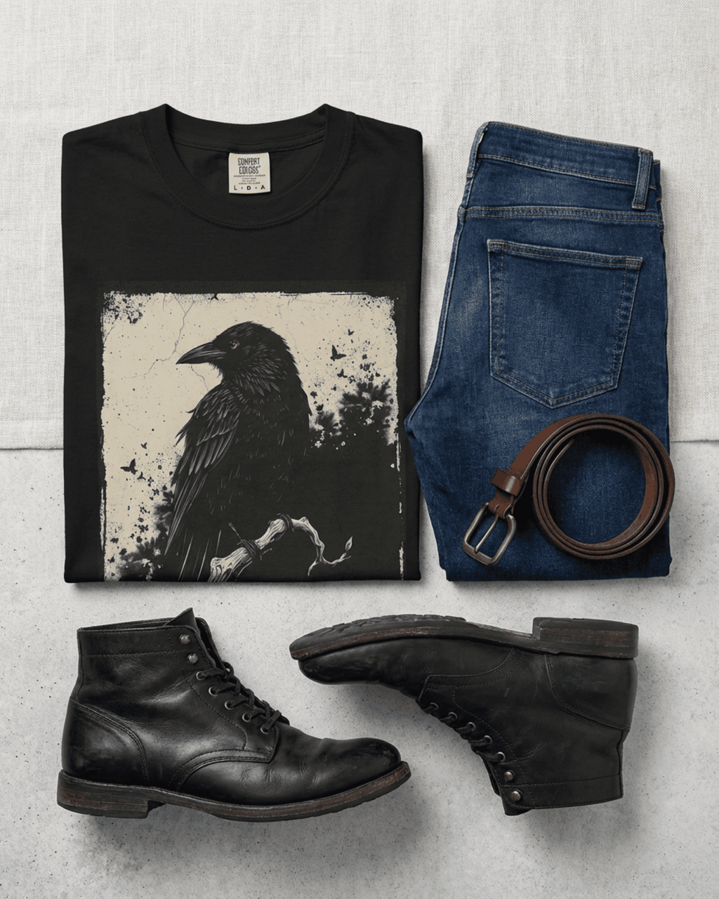 Flat lay of a black Comfort Colors Black Raven Tshirt with a detailed crow illustration, styled with dark blue jeans, a brown leather belt, and worn black boots for a goth alternative style shirt and dark academia aesthetic tee look.