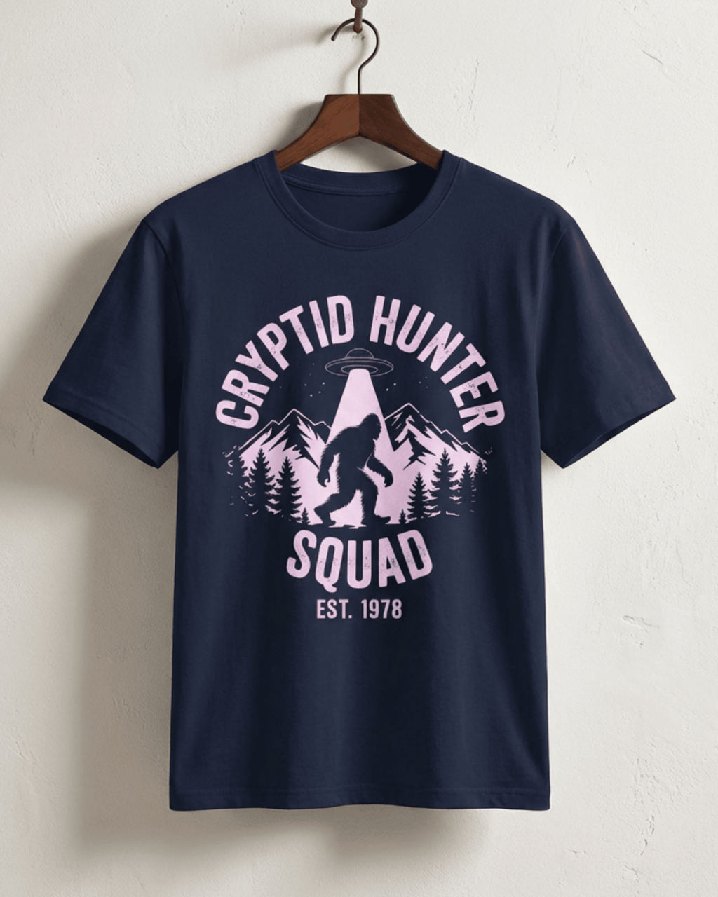The navy Bigfoot Cryptid Hunter T-Shirt hanging on a wooden hanger. This version features the 'Cryptid Hunter Squad' design in a striking off-white print, showing Sasquatch and a UFO, making it a unique paranormal-themed gift for men and women.