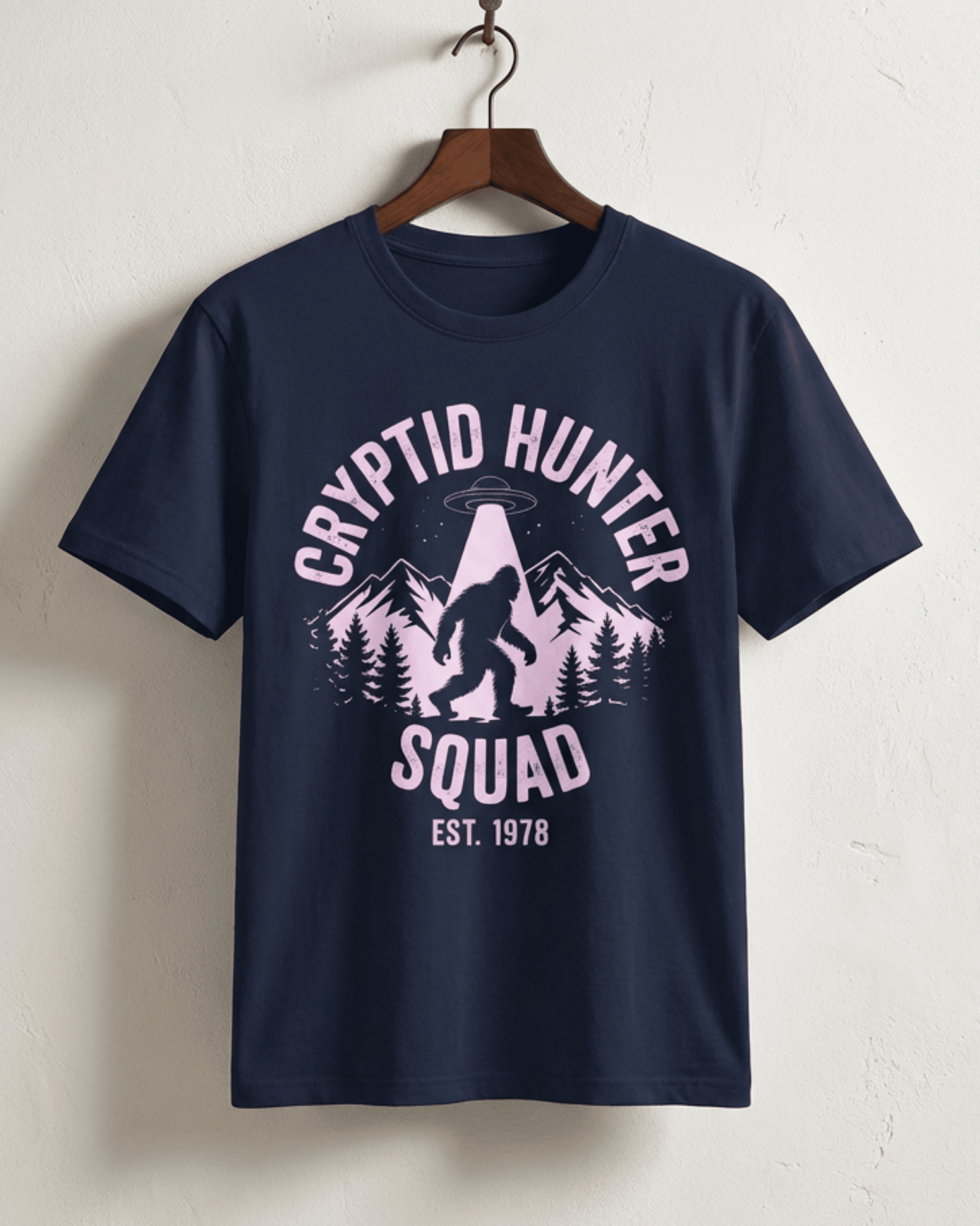 The navy Bigfoot Cryptid Hunter T-Shirt hanging on a wooden hanger. This version features the 'Cryptid Hunter Squad' design in a striking off-white print, showing Sasquatch and a UFO, making it a unique paranormal-themed gift for men and women.