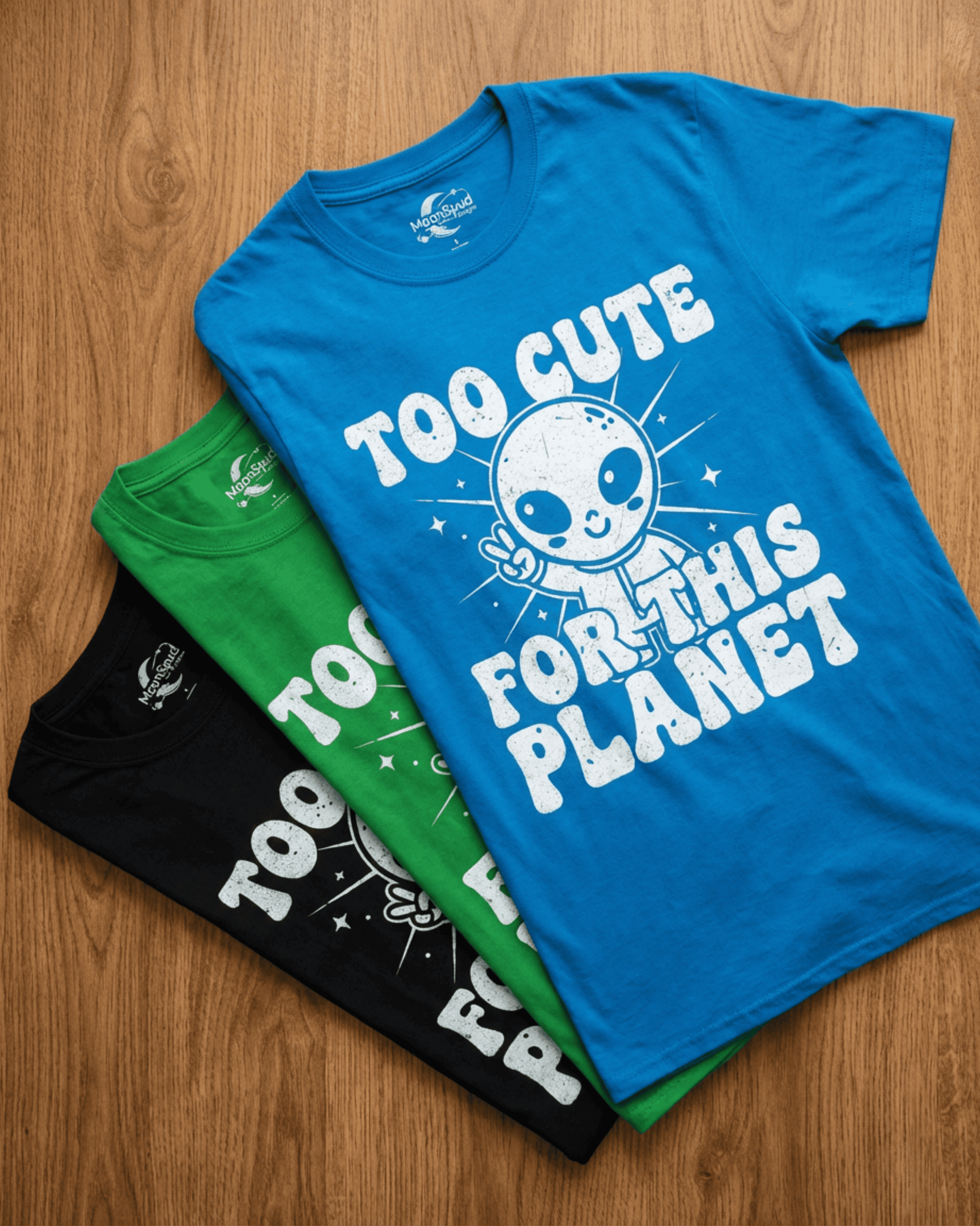 A flat lay of our funny graphic tee for outer space lovers, showing the cute alien peace sign shirt in blue, green, and black. The retro 90s Y2K style design says "Too Cute For This Planet" in a bubbly white font.