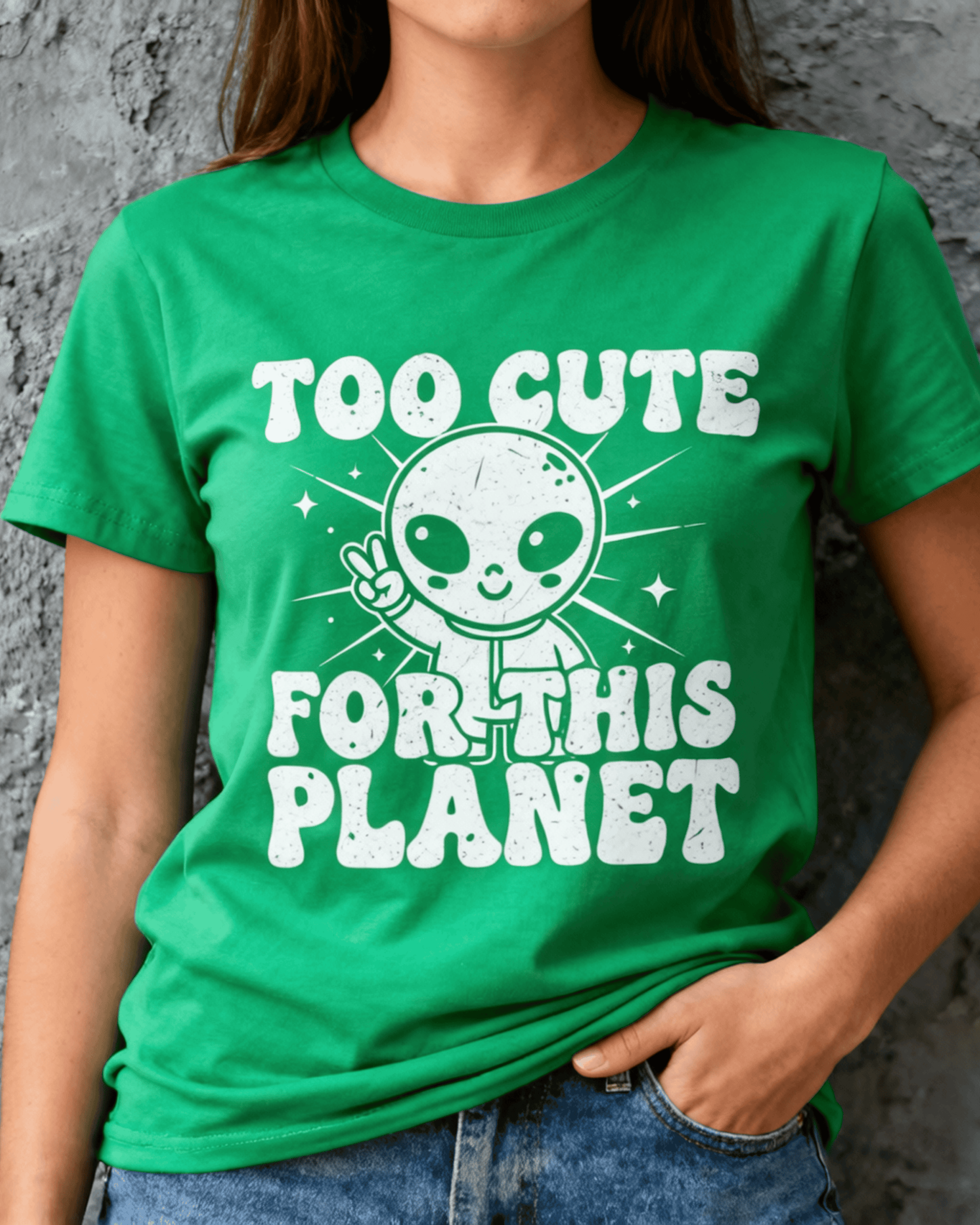 Close-up on the chest of a woman wearing a green Cute Alien Peace Sign T-Shirt. The funny graphic tee features a white retro print of an alien with the text 'Too Cute For This Planet' against a plain grey wall.
