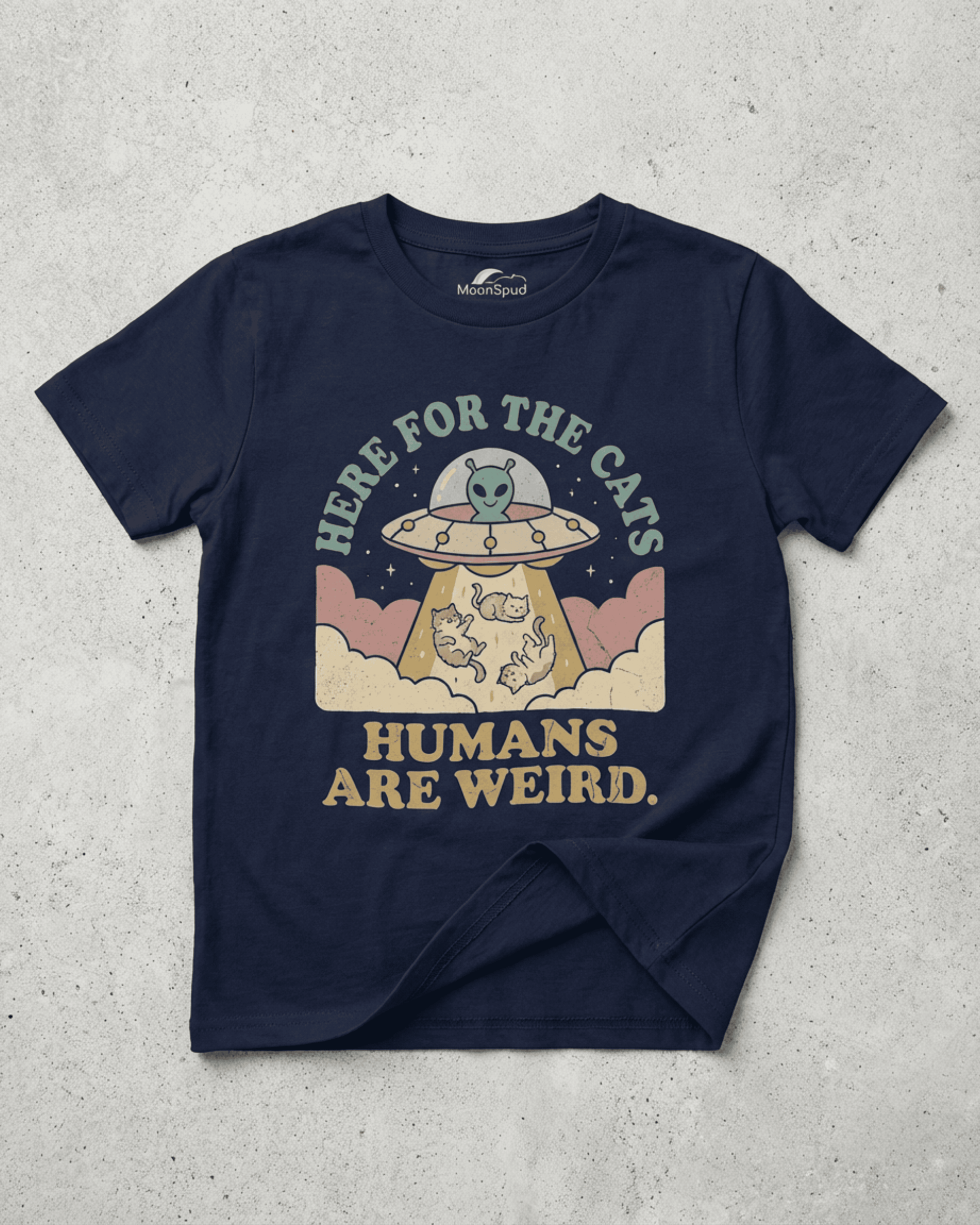 A navy blue Cute Kids Alien Cat T-Shirt laid flat on a textured surface. The graphic clearly shows a UFO, a smiling alien, and three cats in a tractor beam with the funny phrases "Here for the Cats" and "Humans Are Weird."