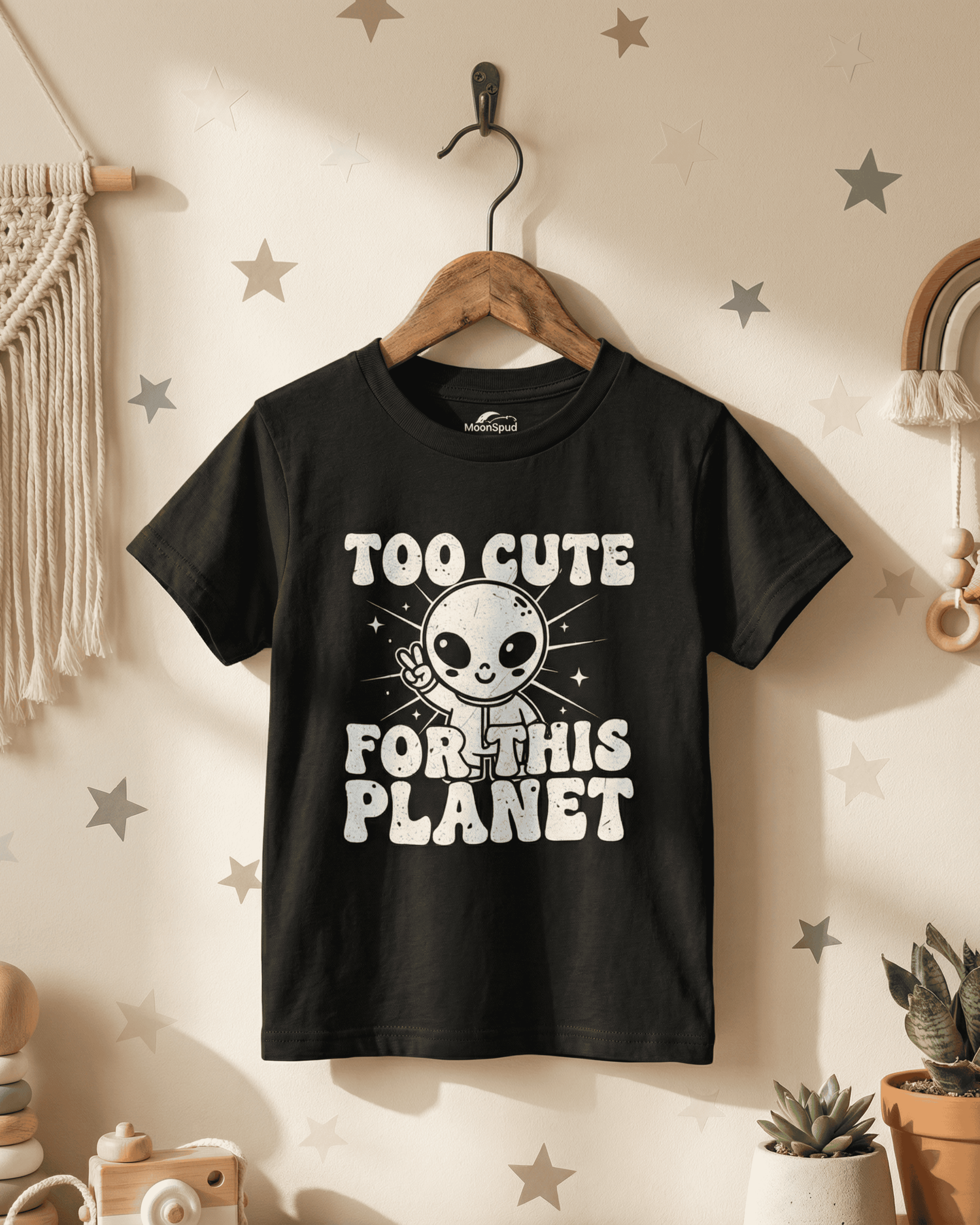 The cute kids alien tshirt in black hangs on a wooden hanger in a stylish child's room. This piece of youth space-themed apparel shows off the "Too Cute For This Planet" design in a real-life setting, perfect for any kid's wardrobe.