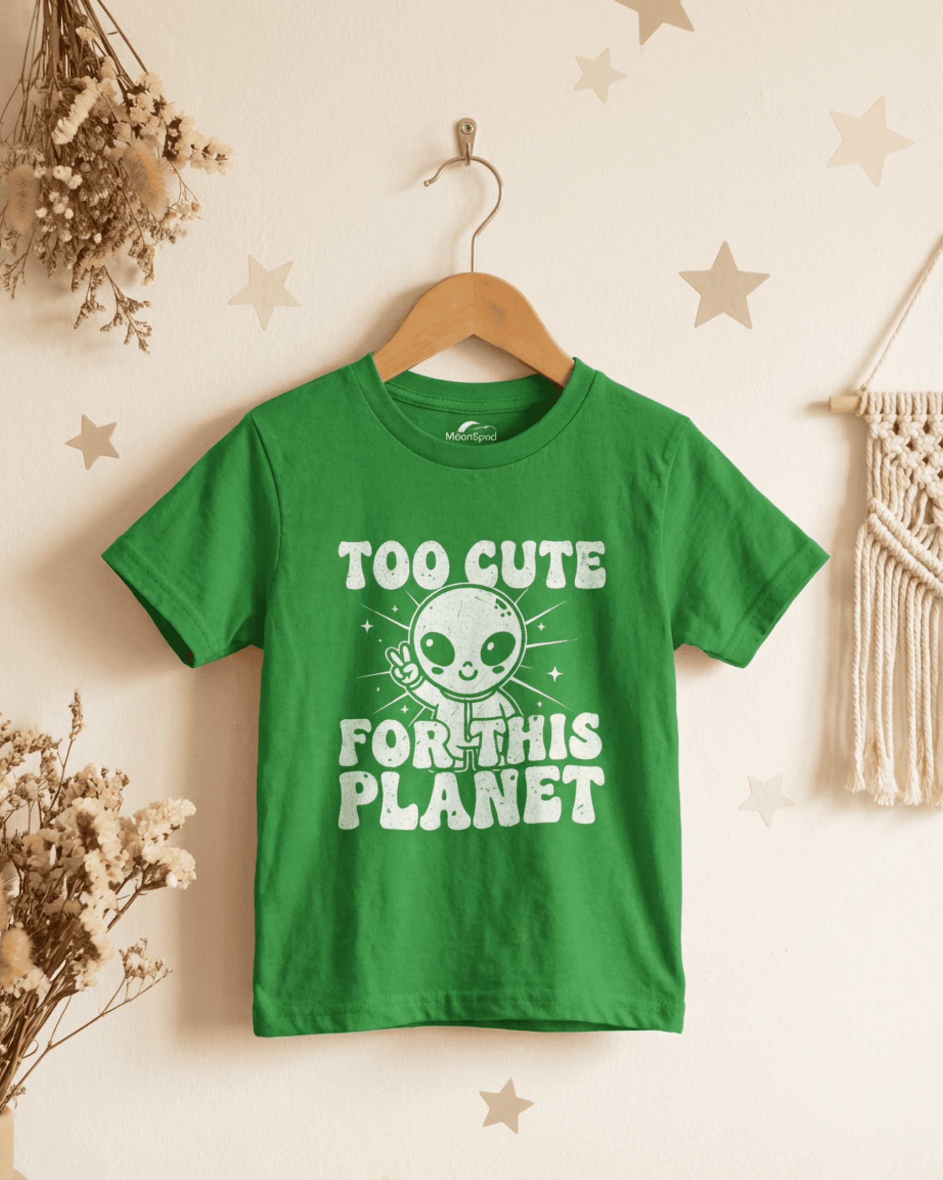 A detail shot of the kelly green version of the cute kids alien tshirt hanging from a hook. This playful toddler or youth shirt is shown in a warmly lit room, highlighting its fun extraterrestrial design and soft cotton material.