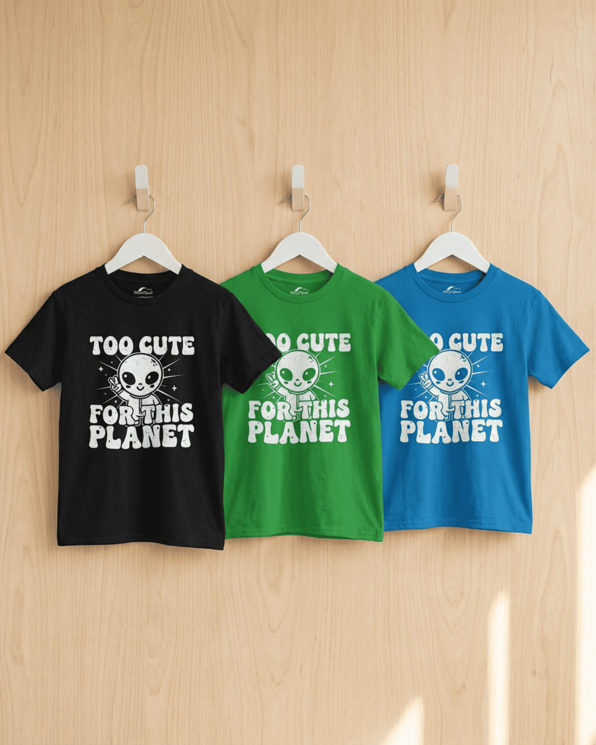 Three cute kids alien tshirts in black, green, and blue hanging side-by-side from white hangers on a light wood-paneled wall. Each youth graphic tee features the "Too Cute For This Planet" design, showcasing the available color options.