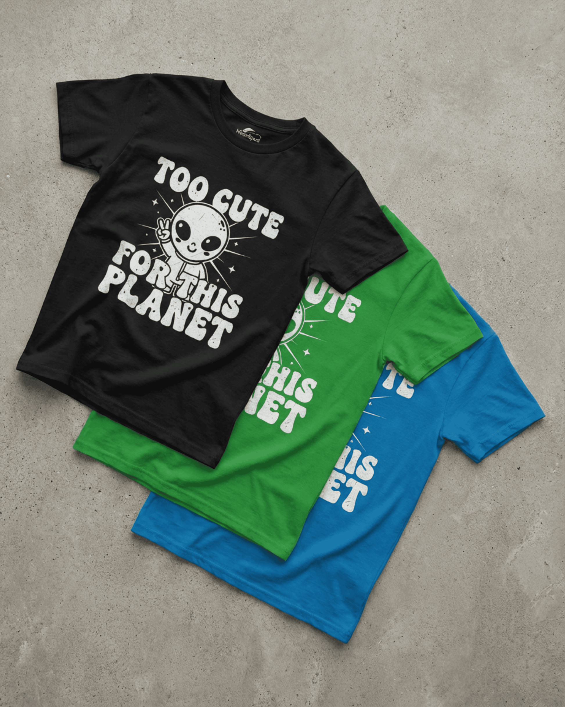 A black version of the cute kids alien tshirt is laid flat, displaying the full design of a cartoon alien giving a peace sign with the funny text "Too Cute For This Planet." The green and blue shirt options are visible underneath.