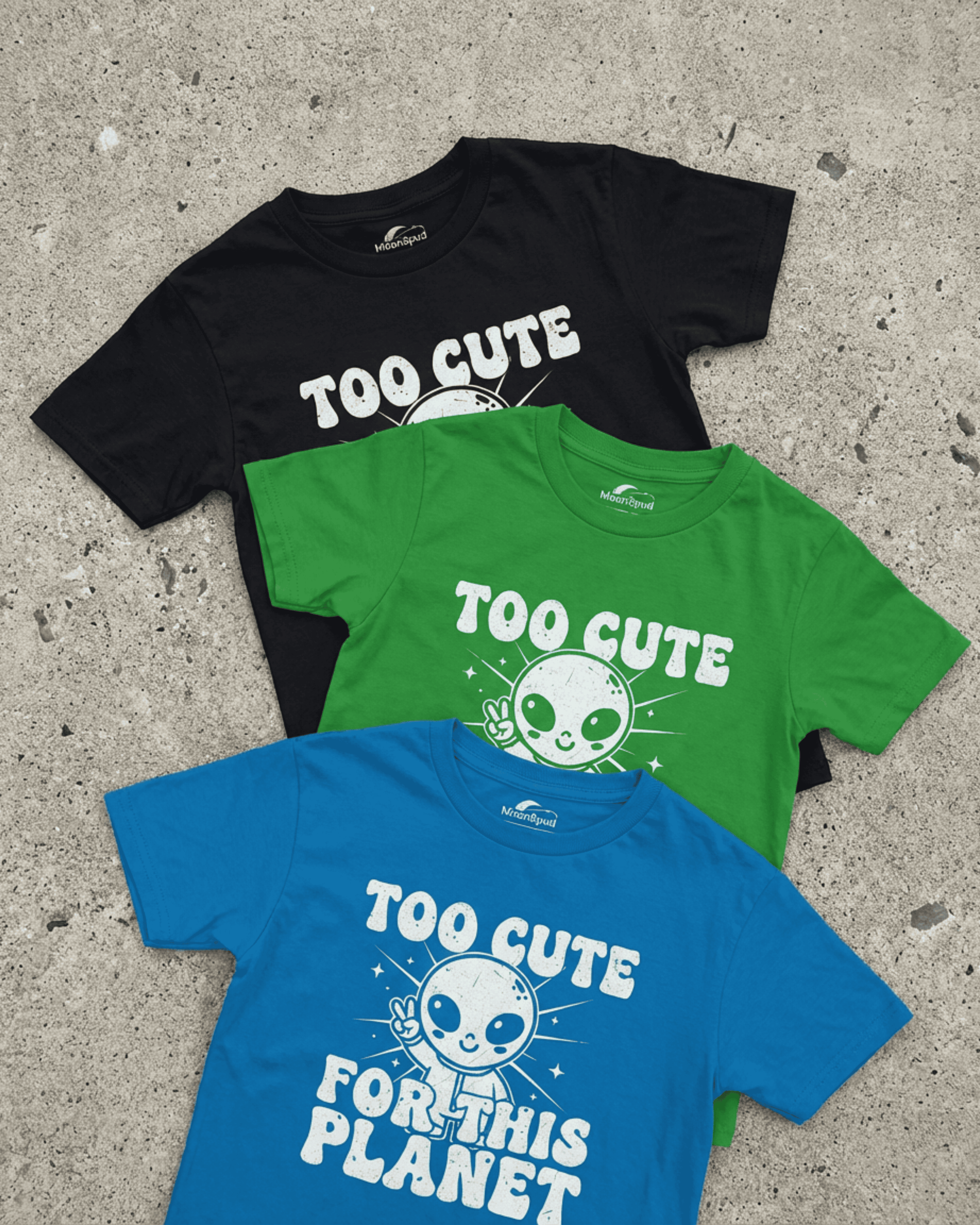 A flat lay of three cute kids alien tshirts on a stone background. A bright blue shirt is in the foreground, showing a friendly alien and the text "Too Cute For This Planet." A green and a black version of the funny kids shirt are layered behind it.