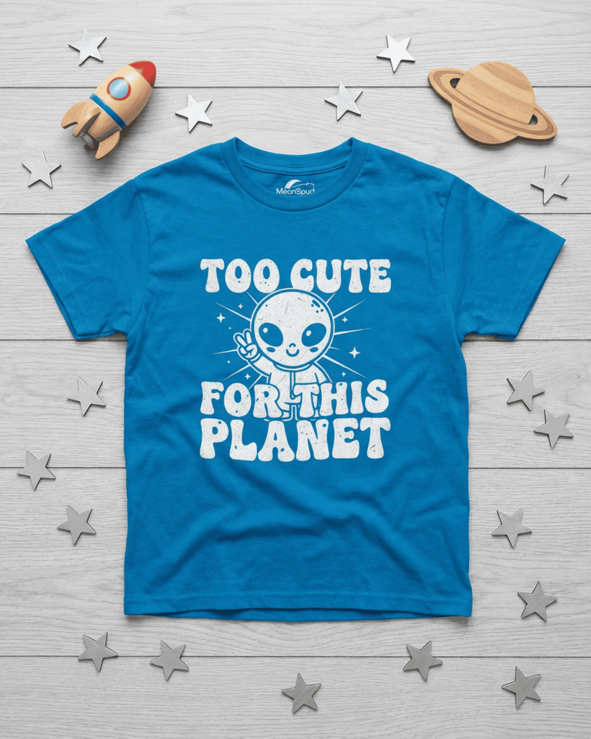 A product shot showing the bright blue color option for our cute kids alien tshirt. This top-down view of the funny children's clothing item displays the "Too Cute For This Planet" graphic alongside a toy rocket and stars, emphasizing the fun, cosmic theme.