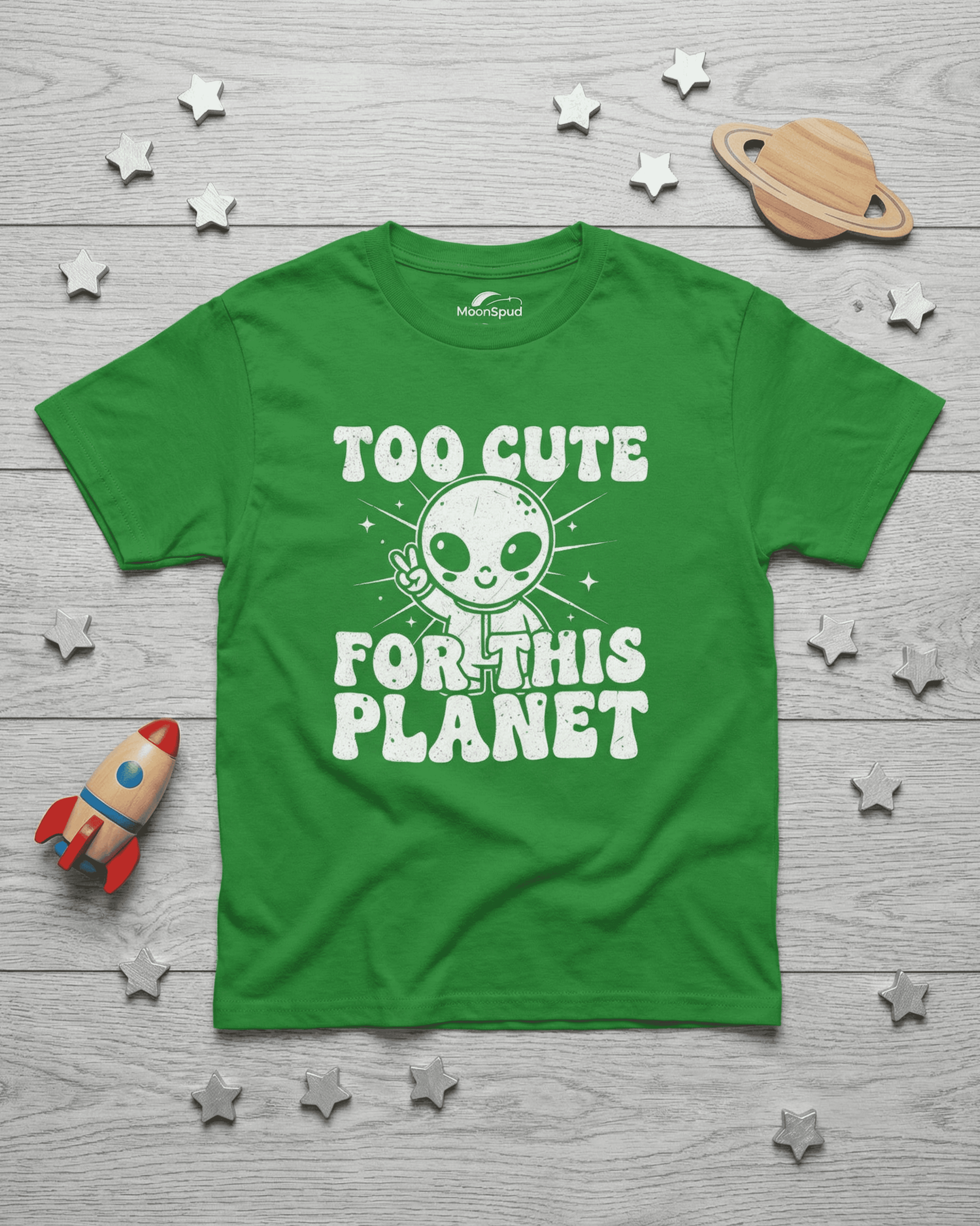A vibrant green cute kids alien tshirt laid flat on a grey wooden surface. The white graphic design, featuring a friendly alien and the words "Too Cute For This Planet," stands out on this unique children's outer space top.