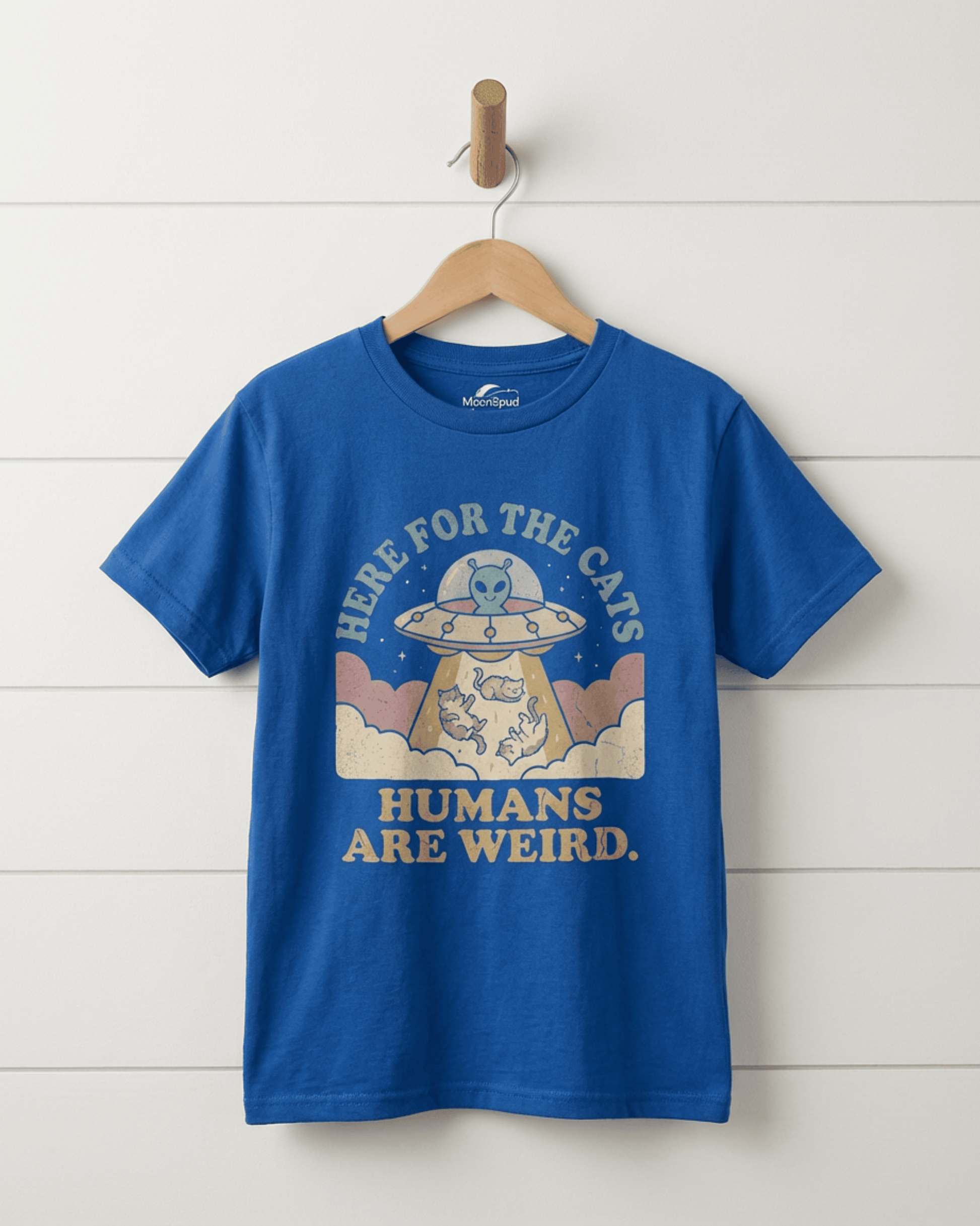 The royal blue version of the Cute Kids Alien Cat T-Shirt hanging against a white wall. The "UFO Here for the Cats" design is clearly visible, making it a fun "Humans Are Weird" graphic tee.