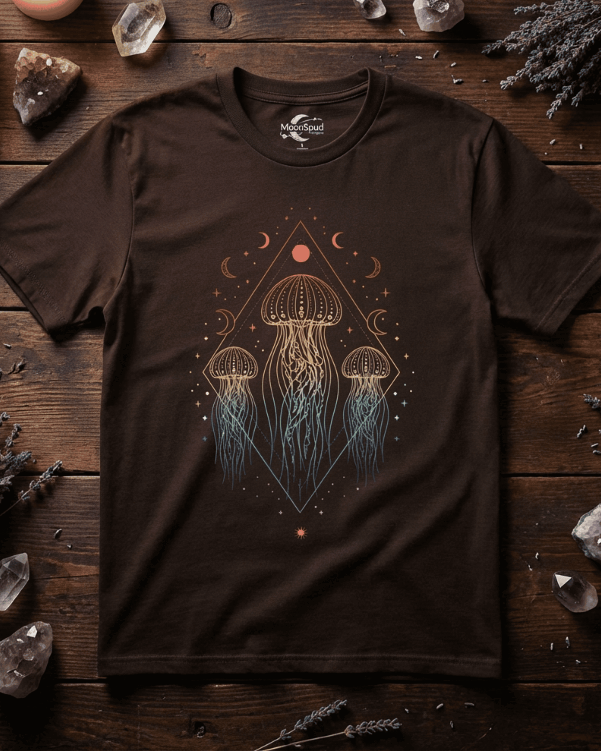 A dark brown Jellyfish Rainbow Festival Tshirt styled in a flat lay on a dark wood background. The celestial boho shirt is surrounded by amethyst crystals and lavender, highlighting the intricate graphic of three ocean jellyfish glowing in a rainbow gradient.