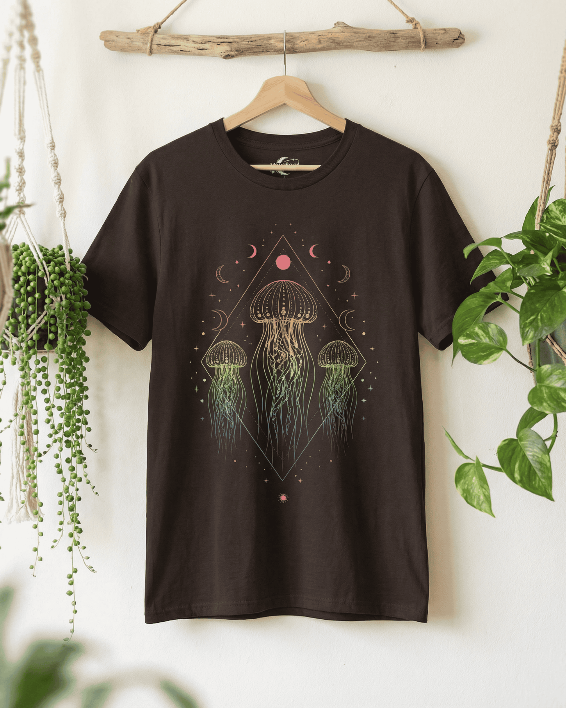 The dark brown Sea Animals Top shown on a wooden hanger against a white wall. This celestial jellyfish t-shirt is displayed between a hanging string-of-pearls plant and a pothos, creating a fresh, bohemian vibe.