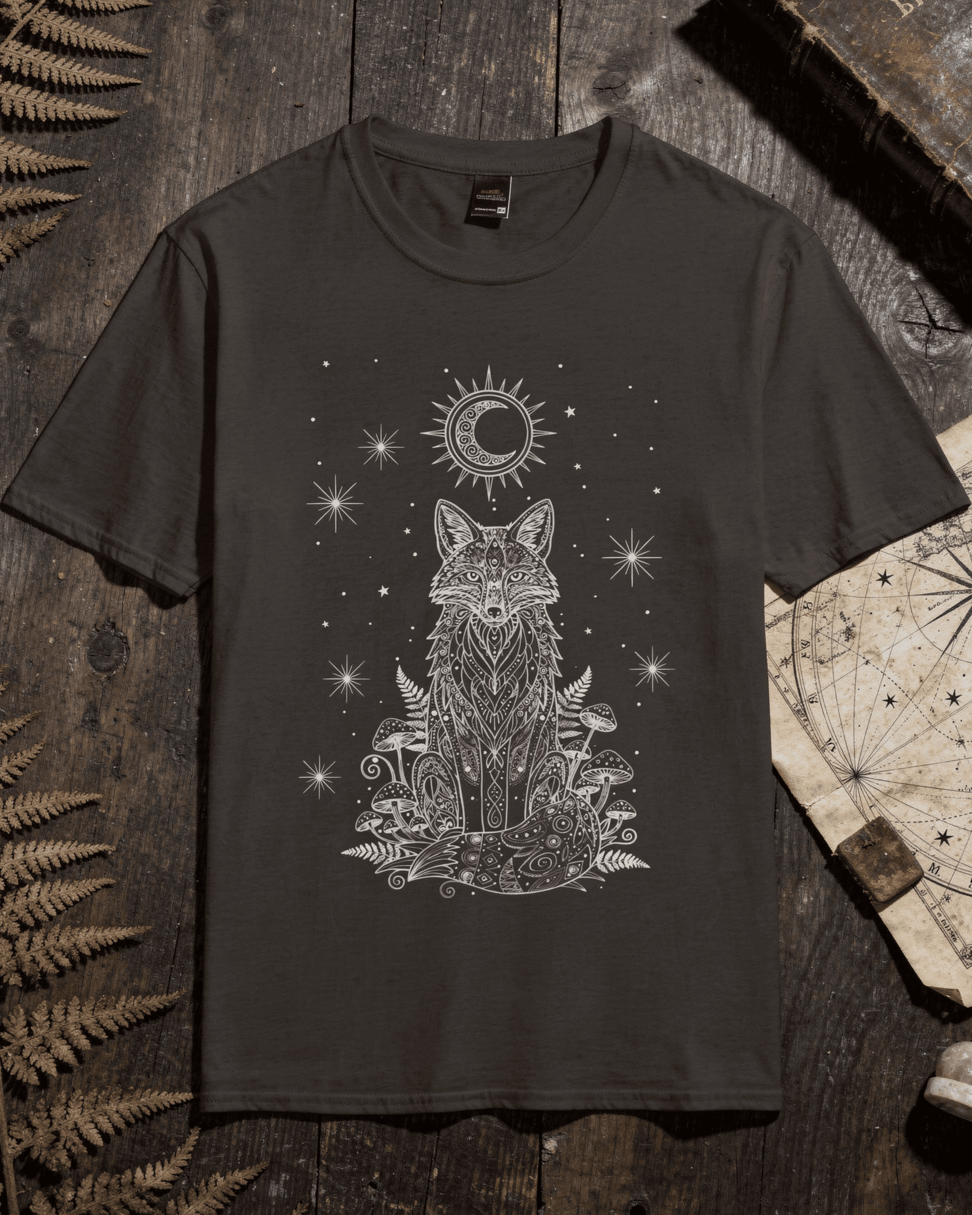 A flat lay top-down view of a dark brown Fox Shirt placed on a rustic wooden table next to dried ferns and an antique map, displaying the Woodland Top design of a sitting fox, celestial stars, and a crescent moon.