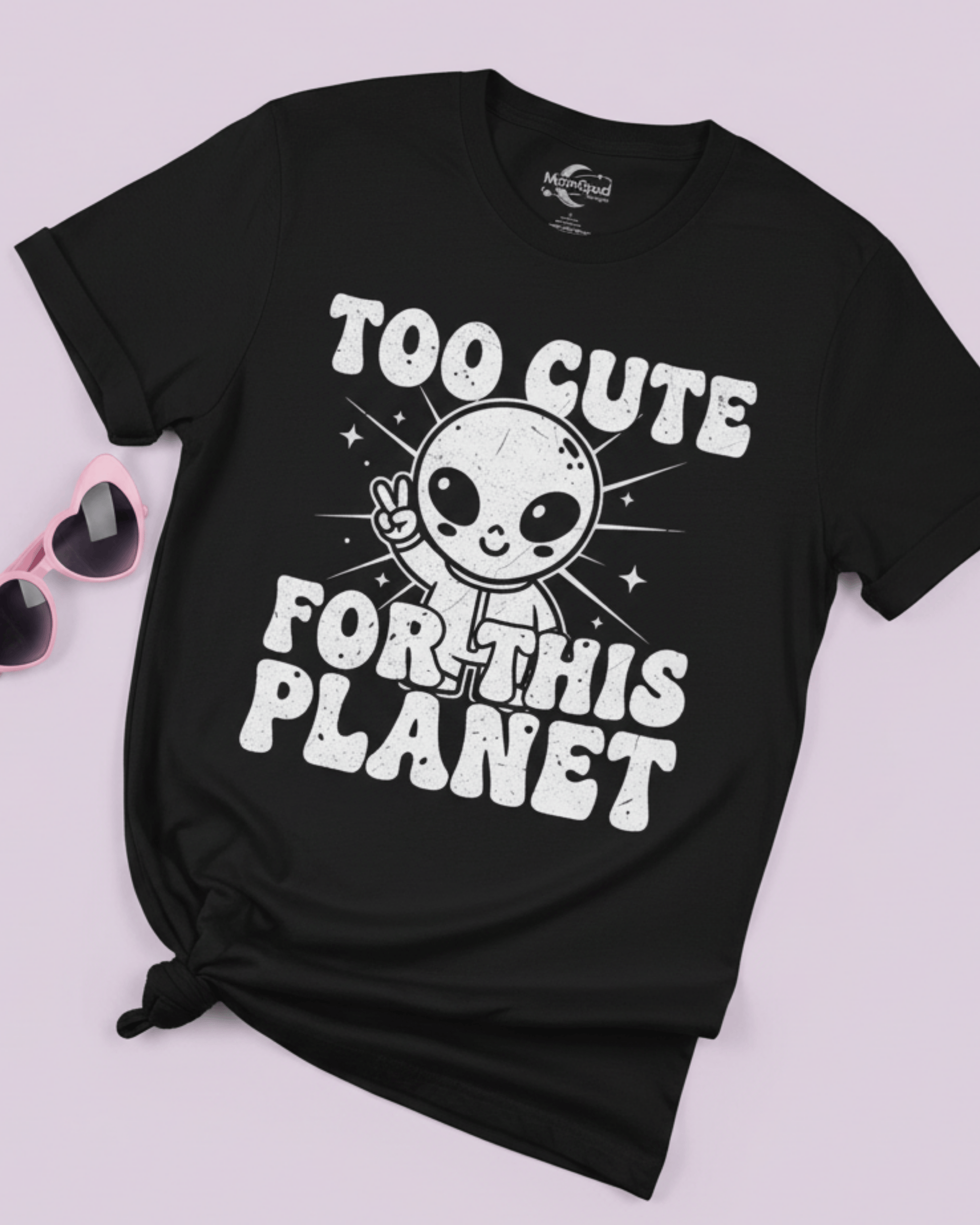 A flat lay of the black funny graphic tee, tied in a knot to showcase a popular Y2K styling option. The cute alien and 'Too Cute For This Planet' design is shown next to pink sunglasses, perfect for teen girls.