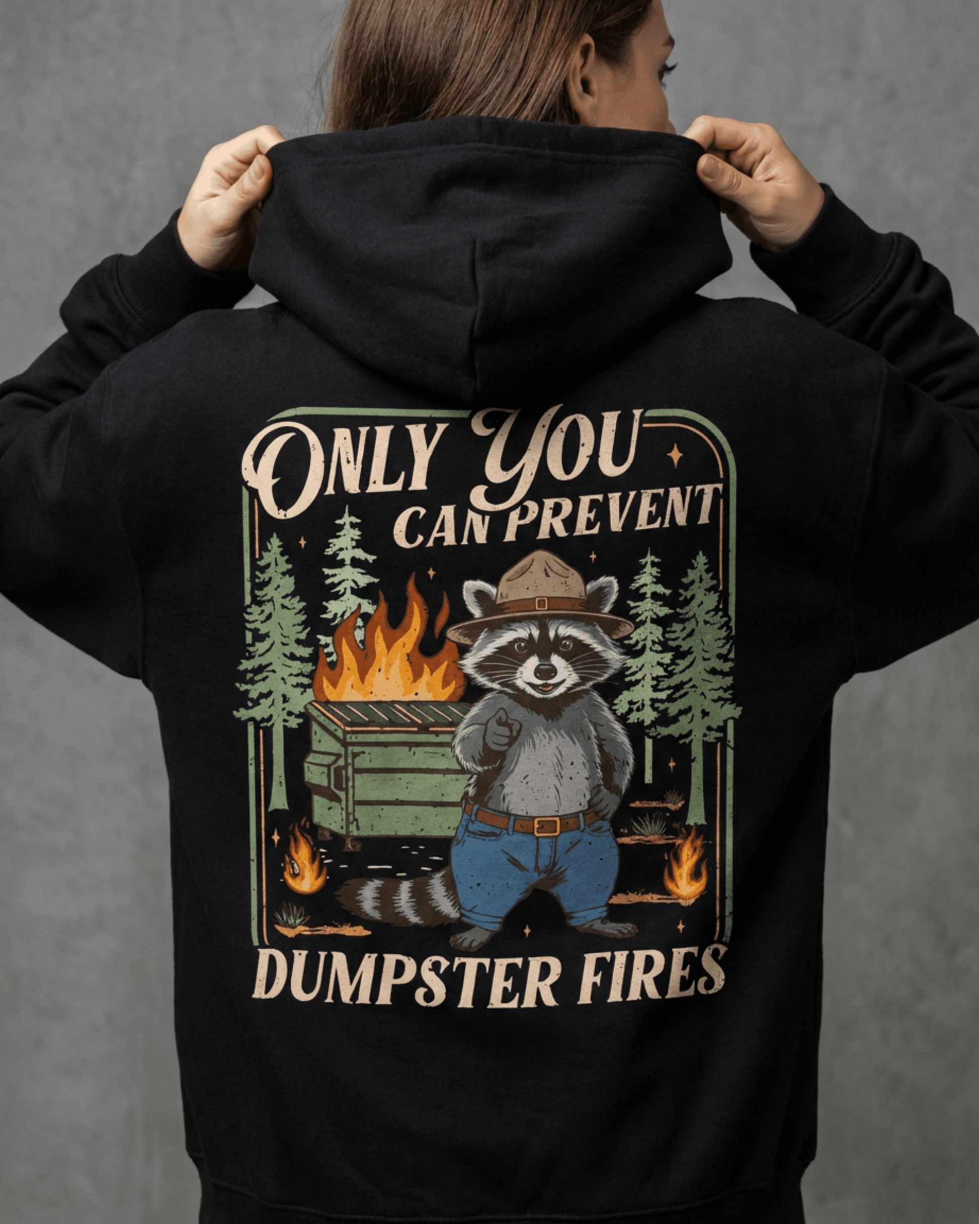 A detailed look at the funny mental health hooded sweatshirt's back design, a National Park parody where a raccoon in a ranger hat says 'Only You Can Prevent Dumpster Fires'.