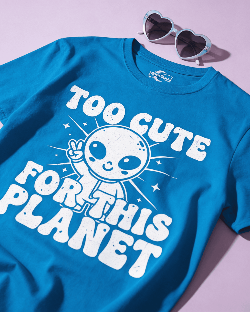 A detailed flat lay of the 'Too Cute For This Planet' Funny Graphic Tee in blue. This UFO Sci Fi Shirt is styled with heart-shaped sunglasses, highlighting the cute alien peace sign design and its fun, retro 90s Y2K vibe.