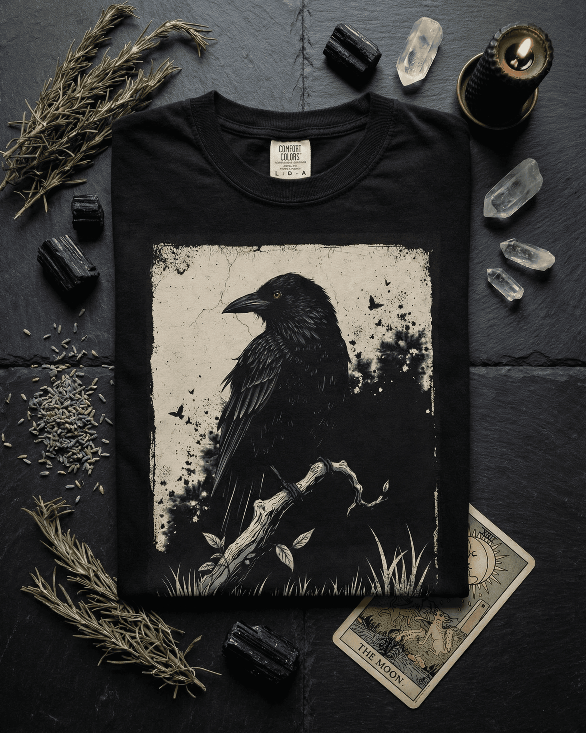 A mystical flat lay of the Goth Alternative Style Shirt featuring a raven graphic. The black tee is arranged on a dark slate surface with black tourmaline and clear quartz crystals, dried herbs, a lit black candle, and The Moon tarot card, perfect for a grunge witchy aesthetic.