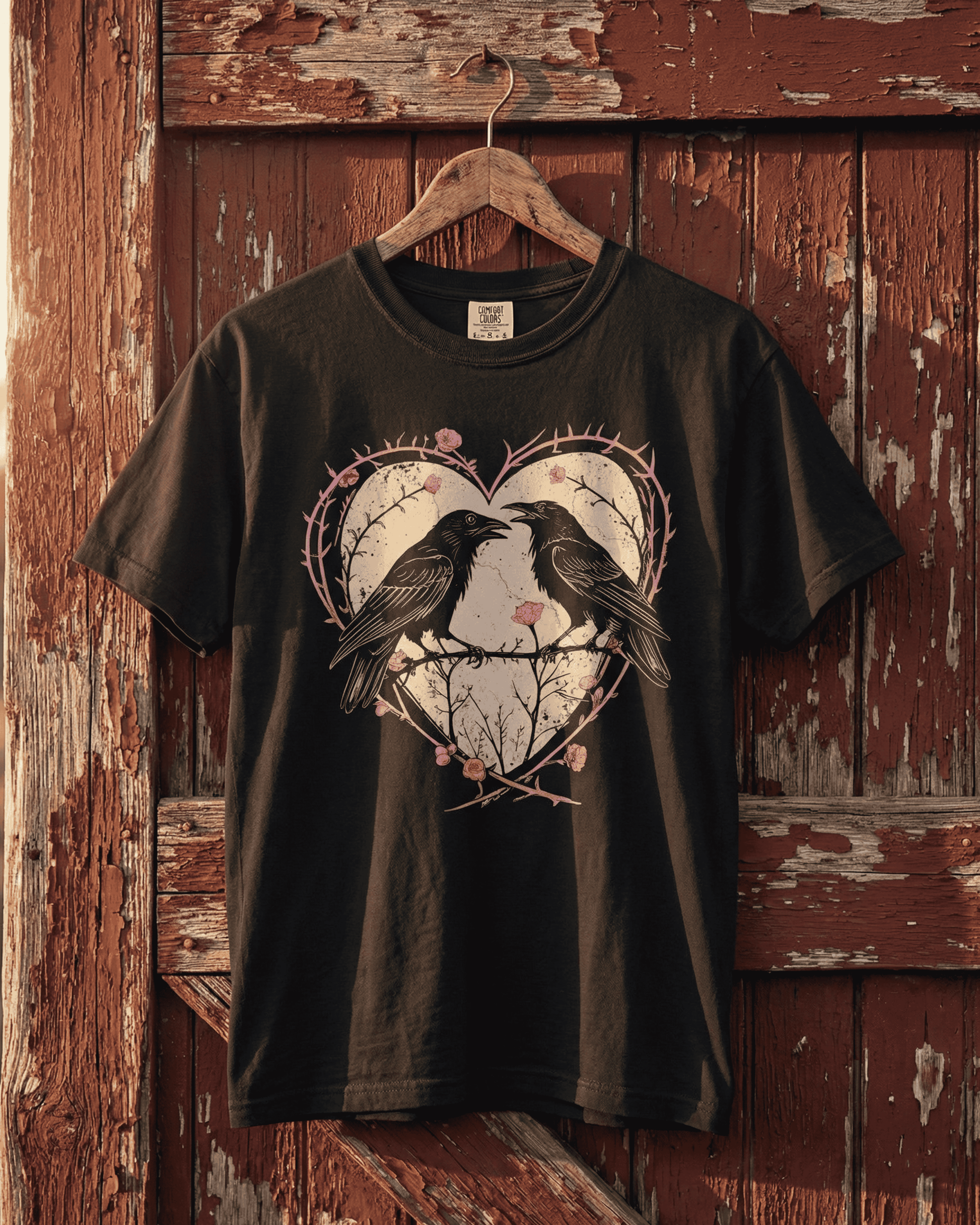 Our black Gothic Dark Romance crow tee hanging against a rustic, weathered red barn door. The edgy punk graphic of two ravens inside a heart of pink thorns and roses stands out, making it the perfect shirt for an Anti Valentine's Day statement.