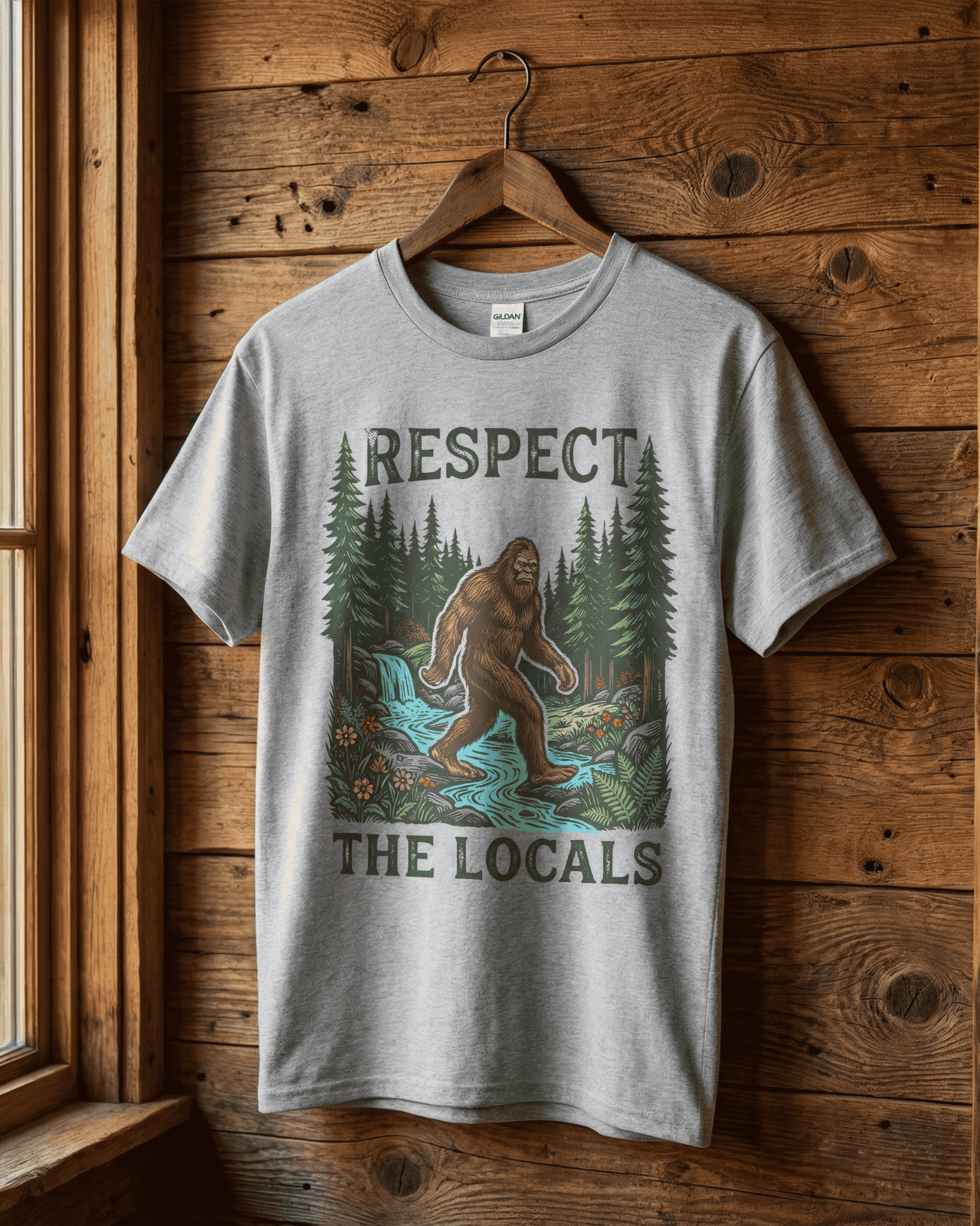 A grey 'Respect the Locals' bigfoot tshirt hanging on a rustic wood wall. The detailed graphic shows the cryptid yeti walking through a forest, making it a great outdoor nature tee for a hiker who loves camping and adventure.