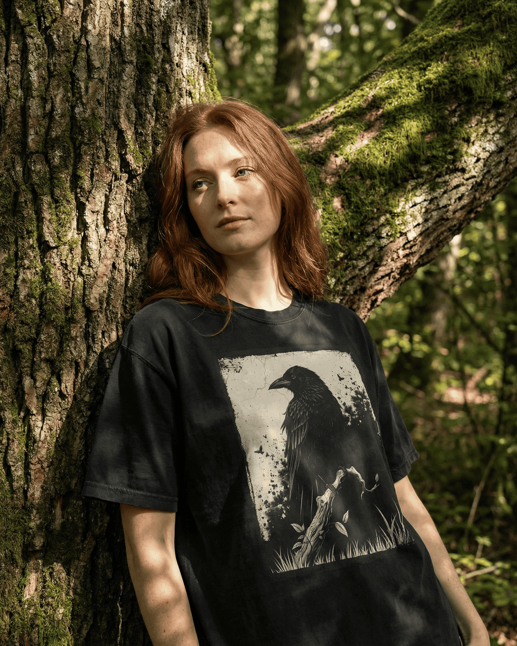 Person standing against a mossy tree in a sunlit forest wearing a black grunge witchy crow t-shirt with a detailed raven and branch design, styled as a nature-inspired dark academia aesthetic tee for women and men.