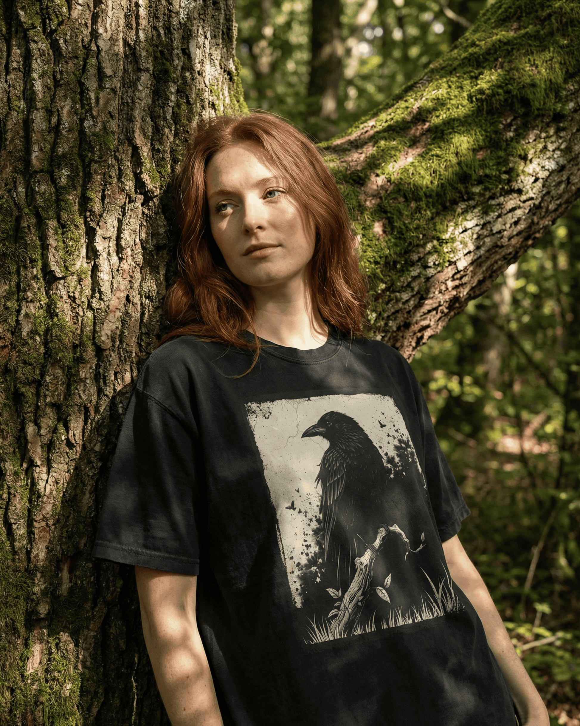 Person standing against a mossy tree in a sunlit forest wearing a black grunge witchy crow t-shirt with a detailed raven and branch design, styled as a nature-inspired dark academia aesthetic tee for women and men.