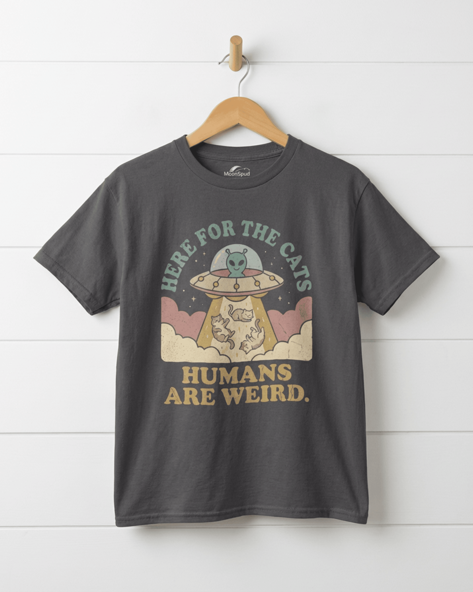 A charcoal "Humans Are Weird" graphic tee for kids hanging on a wooden hanger. The design shows a "UFO Here for the Cats" abducting cats, displayed against a white shiplap wall.