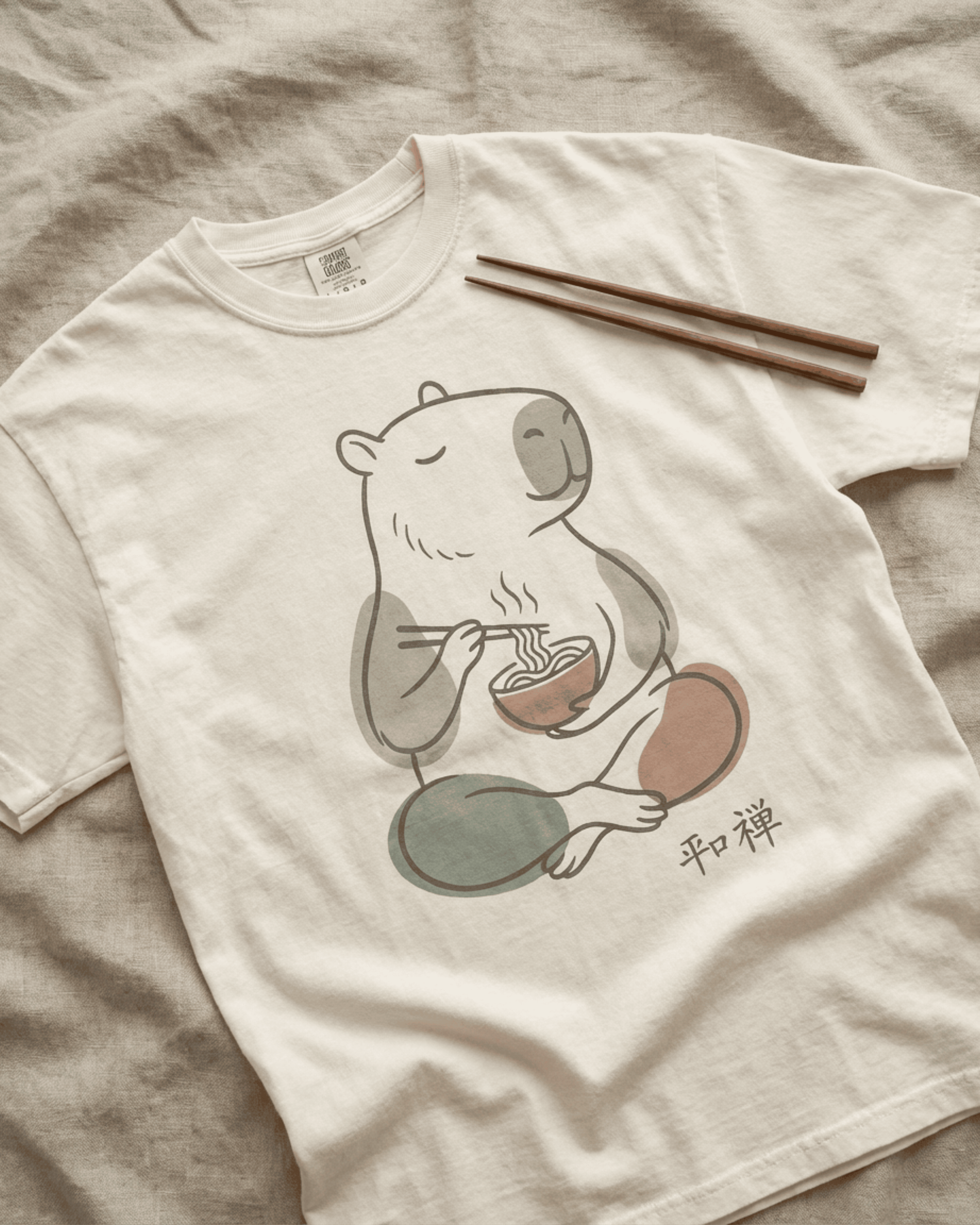A flat-lay image of the Ivory Comfort Colors 1717 tee on a textured linen surface. A pair of wooden chopsticks rests on the shirt, pointing to the serene design of the Capybara Peaceful Zen Ramen T-Shirt, highlighting its relaxed fit and theme.