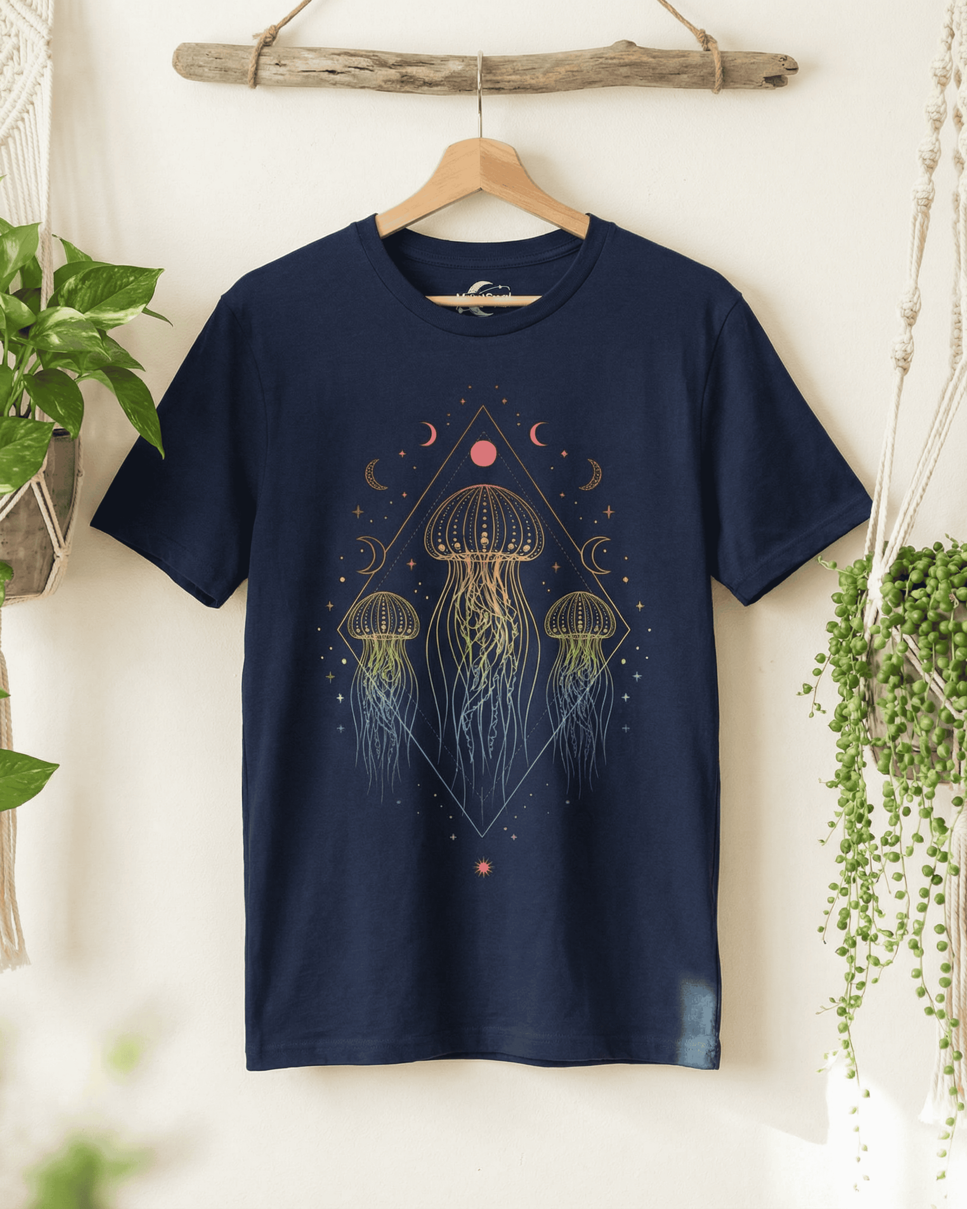 The navy blue Jellyfish Rainbow Festival Tshirt hanging from a piece of weathered driftwood, evoking a perfect ocean lover vibe. This Celestial Boho shirt, with its colorful jellyfish and star design, is flanked by green house plants and macrame wall decor, creating a serene, natural scene.