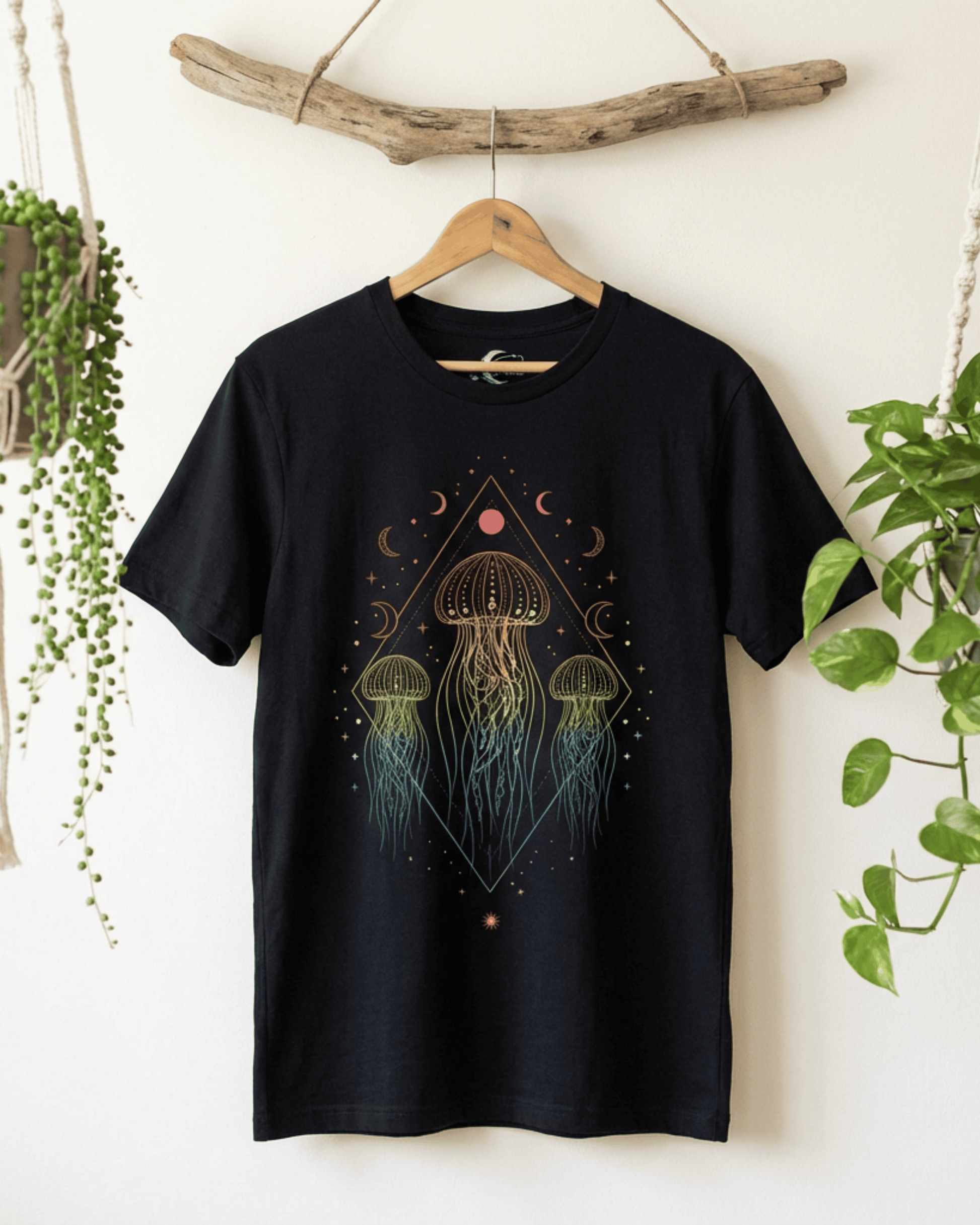 The black Sea Animals Top hanging from a wooden hanger on a piece of driftwood. This celestial boho shirt is styled against a clean white wall with hanging plants, highlighting the detailed rainbow jellyfish and moon graphic.