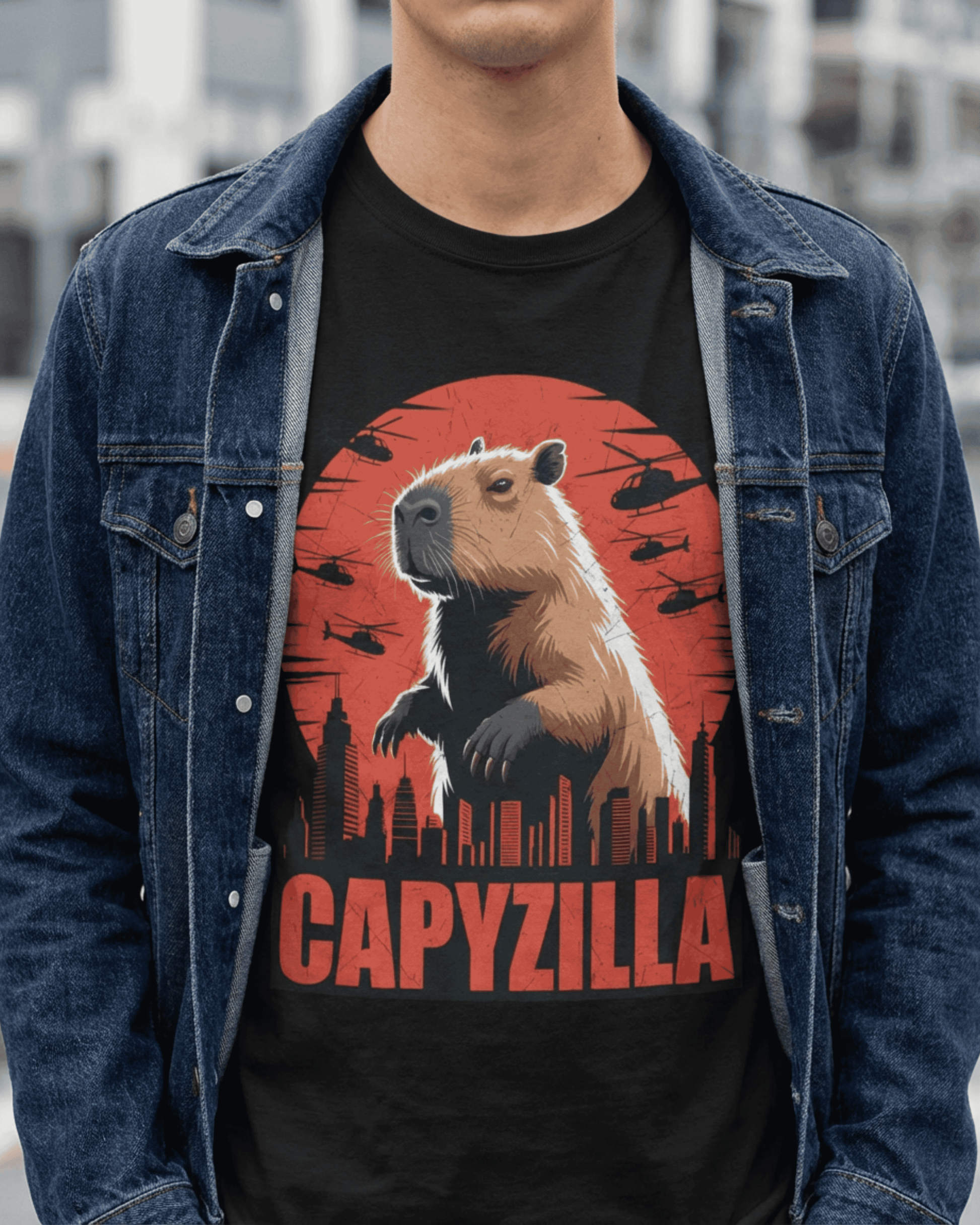 A male model wearing a black Funny Capybara Monster T-Shirt, paired stylishly with a dark blue denim jacket. The prominent 'CAPYZILLA' Godzilla Parody graphic on the front showcases a giant capybara towering over a city, illuminated by a vibrant red sunset, with small helicopters circling. This image highlights the comfortable fit and unique design of the graphic tee on a person.