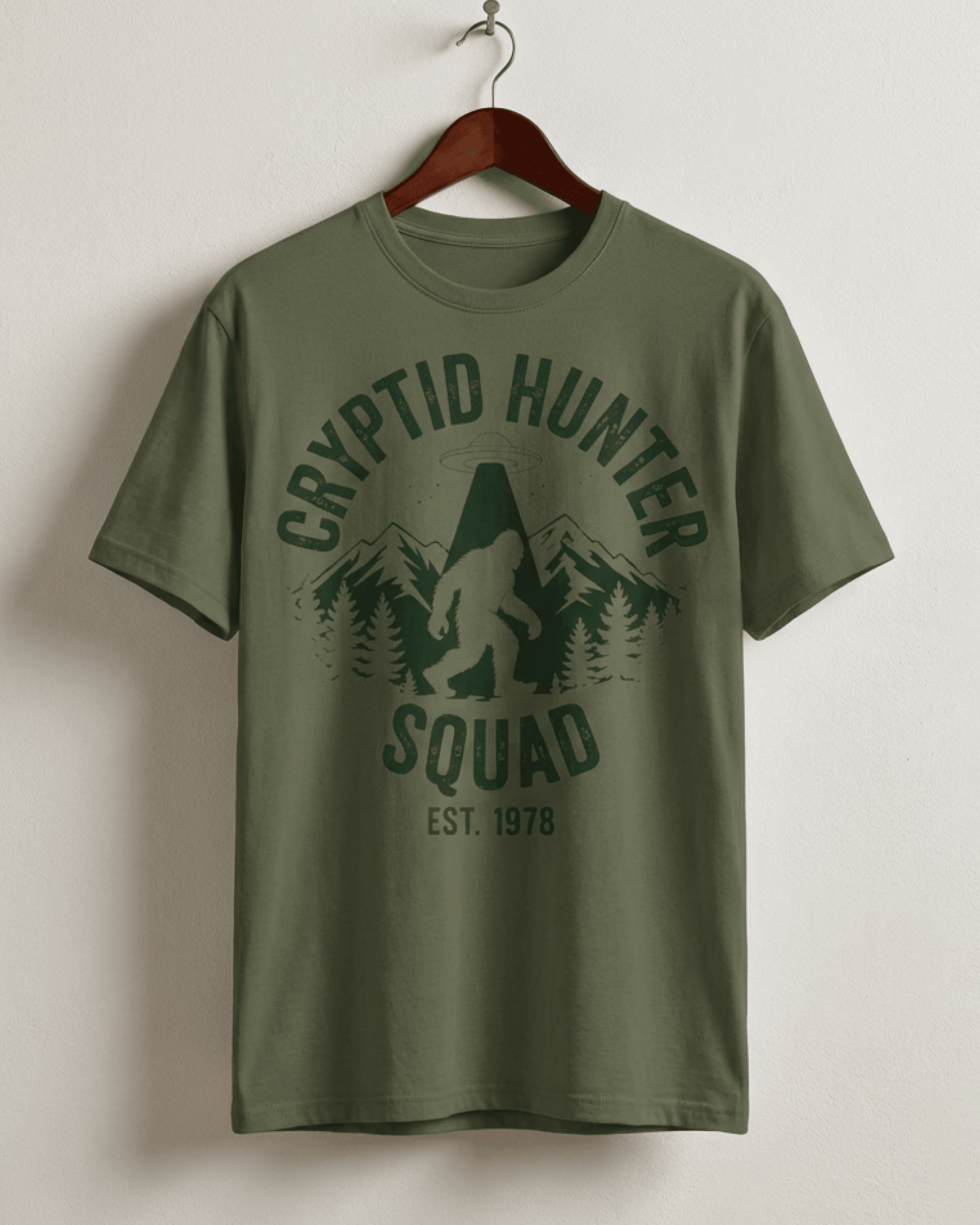 Product shot of the military green Bigfoot Cryptid Hunter T-Shirt on a hanger. This version features a vintage, distressed black print of the iconic 'Cryptid Hunter Squad' design, showing Bigfoot's encounter with a UFO in the wilderness.