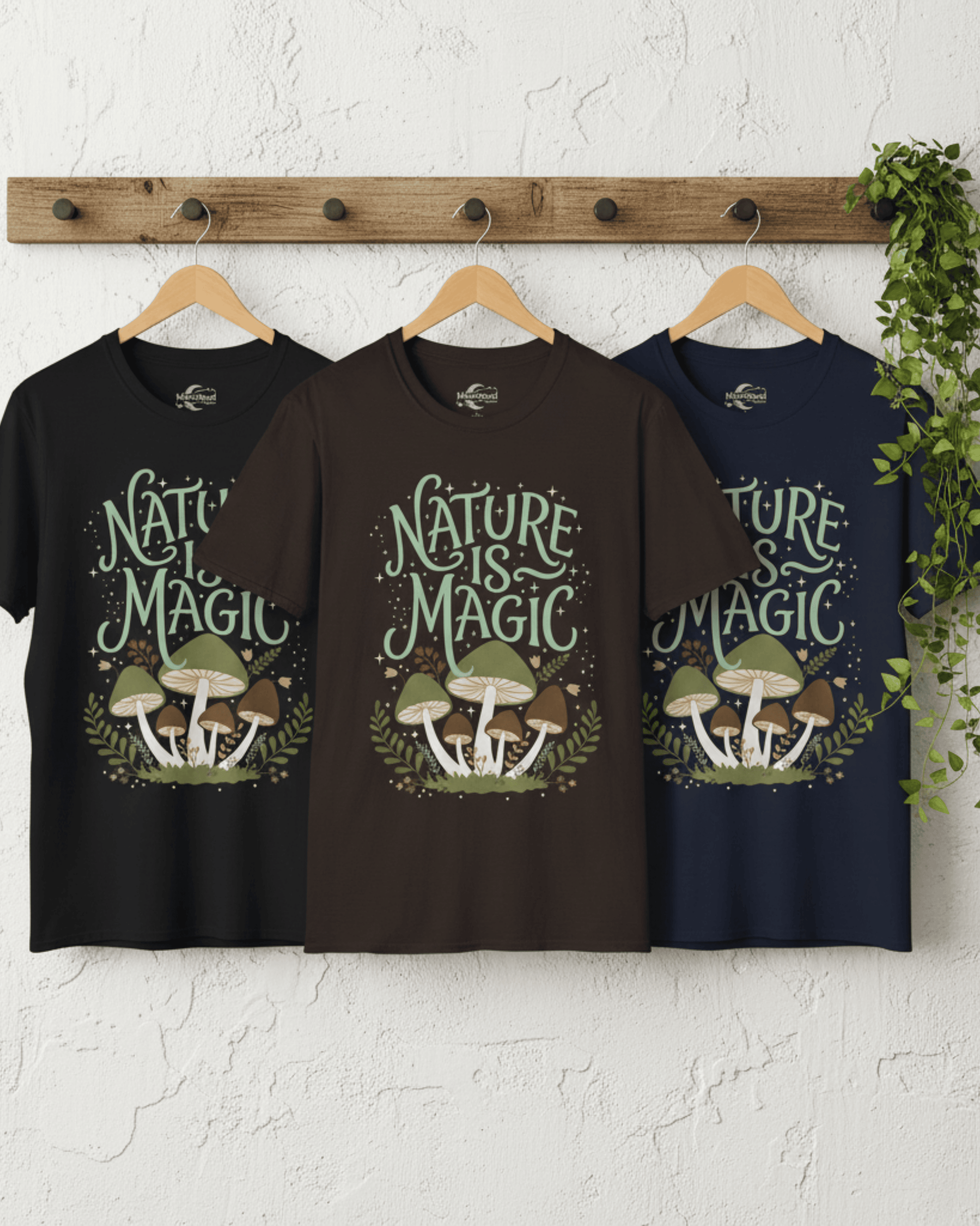 The Nature Is Magic cottagecore t-shirt hanging on wooden hangers in black, dark chocolate, and navy. The detailed botanical mushroom design is clearly visible, showcasing the different color options for this forest-inspired shirt against a clean white wall.