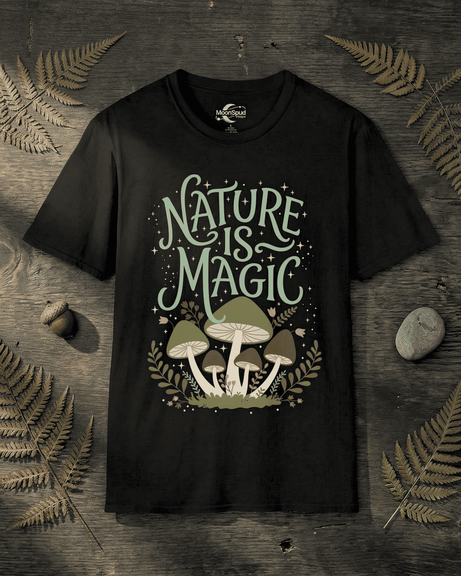 A flat lay of the black Cottagecore Mushrooms Nature Is Magic T-Shirt on a dark, rustic wood background. The shirt's whimsical design features "Nature is Magic" in a light green font above a cluster of enchanting mushrooms and foliage, capturing a magical forest aesthetic.