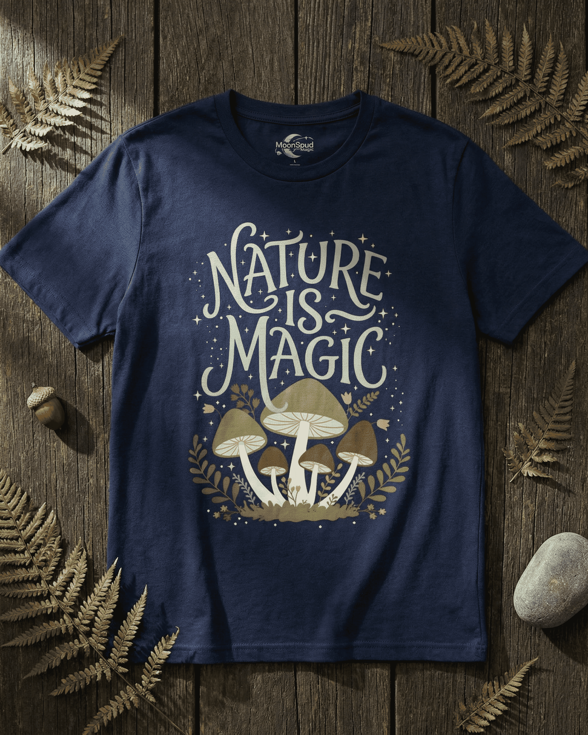 Overhead view of the navy blue Cottagecore Mushrooms Nature Is Magic T-Shirt. The magical design with light green lettering and enchanting mushrooms is showcased on a dark wood table surrounded by dried ferns.