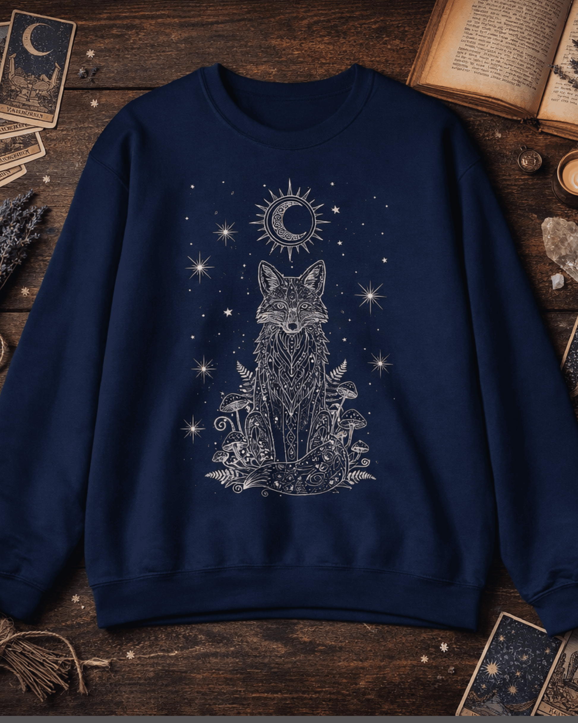 A flat lay of the navy Fox Celestial Moon Sweatshirt, styled with witchy aesthetic props like tarot cards and lavender on a dark wood surface. The mystical crewneck pullover features an intricate silver-white print of a celestial fox sitting amongst mushrooms and ferns under a starry night sky with a sun and crescent moon.