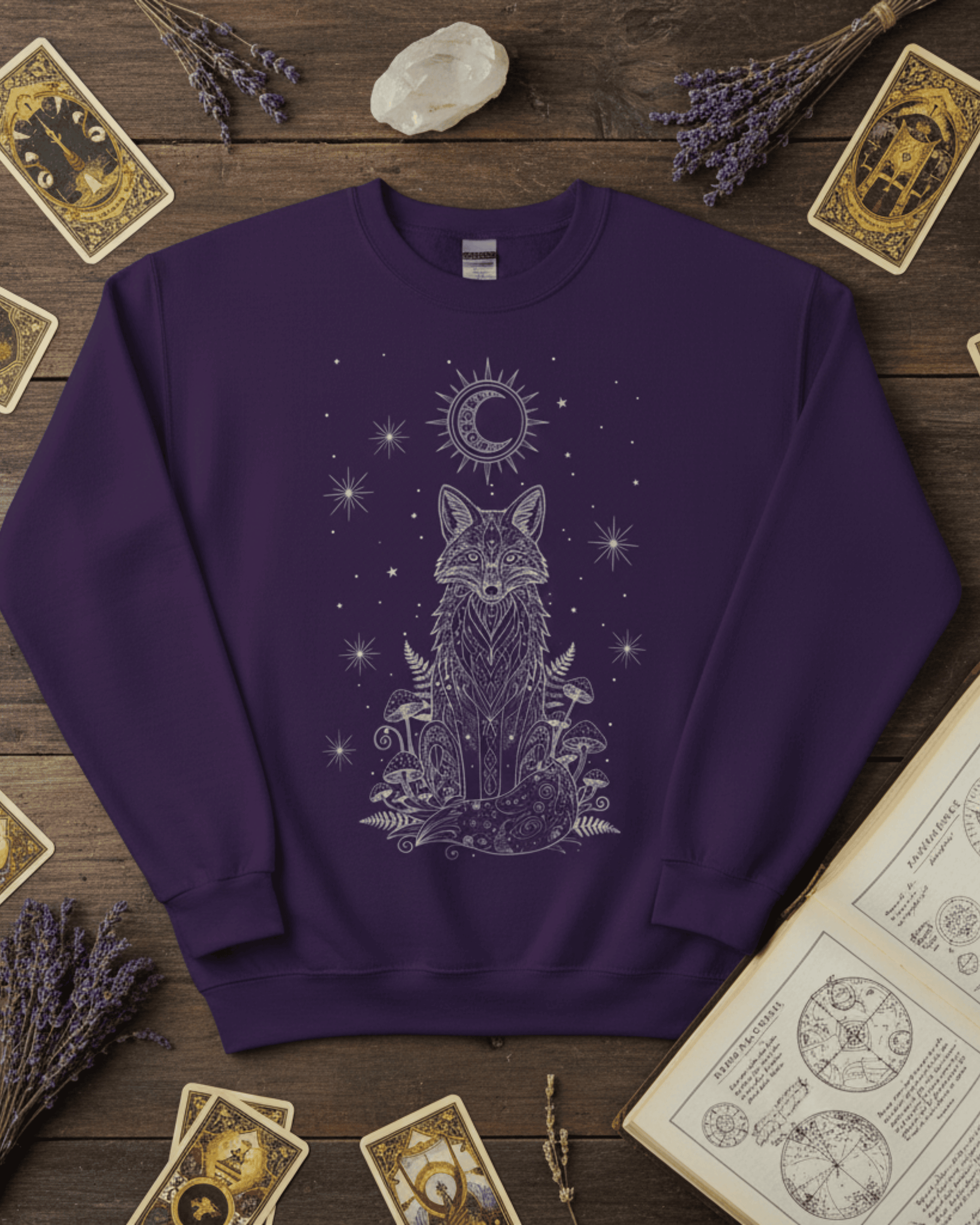 A flat lay of the purple Fox Celestial Moon Sweatshirt, styled on a dark wood background with tarot cards and lavender to evoke a witchy aesthetic. The intricate silver design on this mystical crewneck pullover features a fox sitting amongst mushrooms under a starry sky.