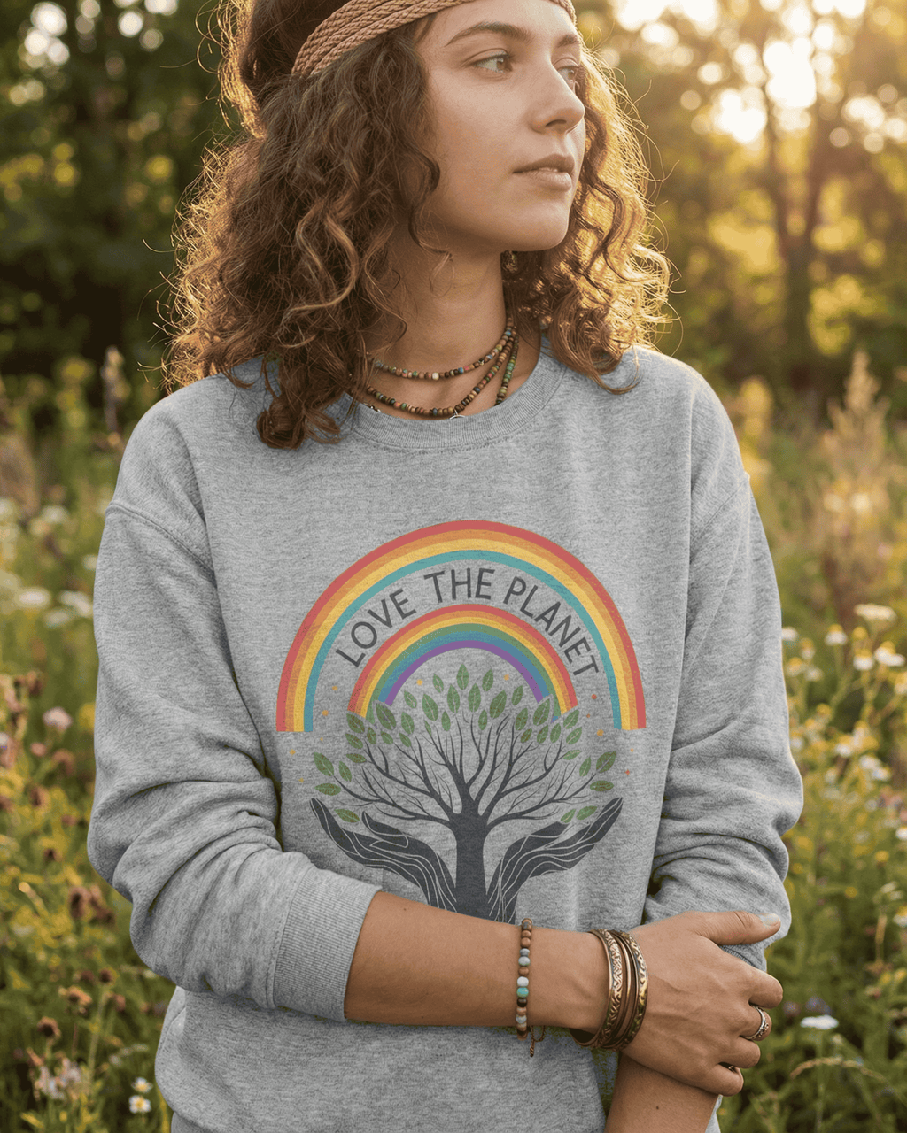 A woman with curly hair stands in a sunny wildflower field wearing the Sport Grey Rainbow Tree of Life pullover. The cozy crewneck displays the "Love The Planet" text within a colorful rainbow, sending a positive message about protecting our environment.