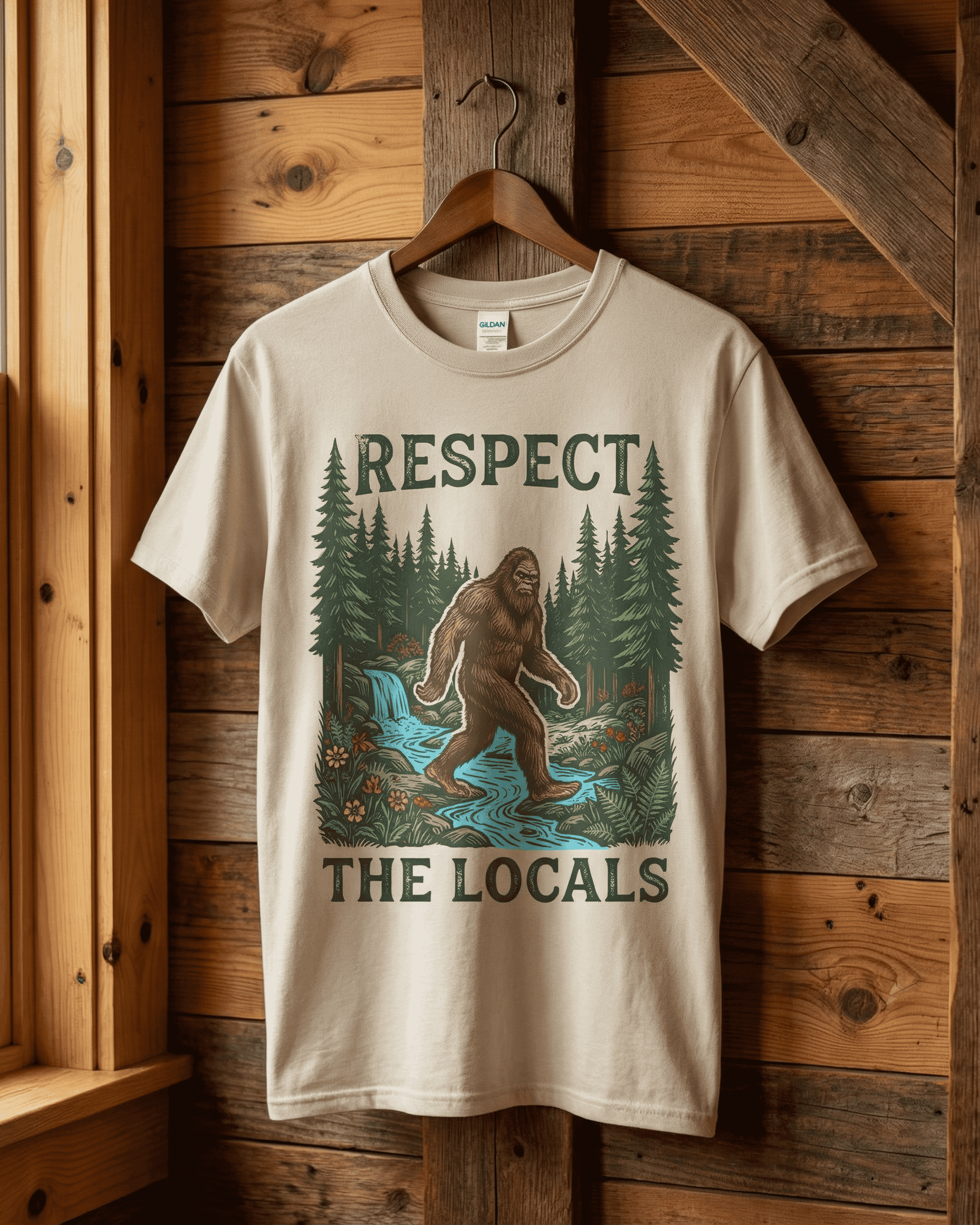 Close-up of the sand beige vintage bigfoot tshirt hanging against a wood panel wall. The 'Respect the Locals' nature tee shows the cryptid walking in a lush forest, a perfect retro shirt for hikers and fans of the outdoors.