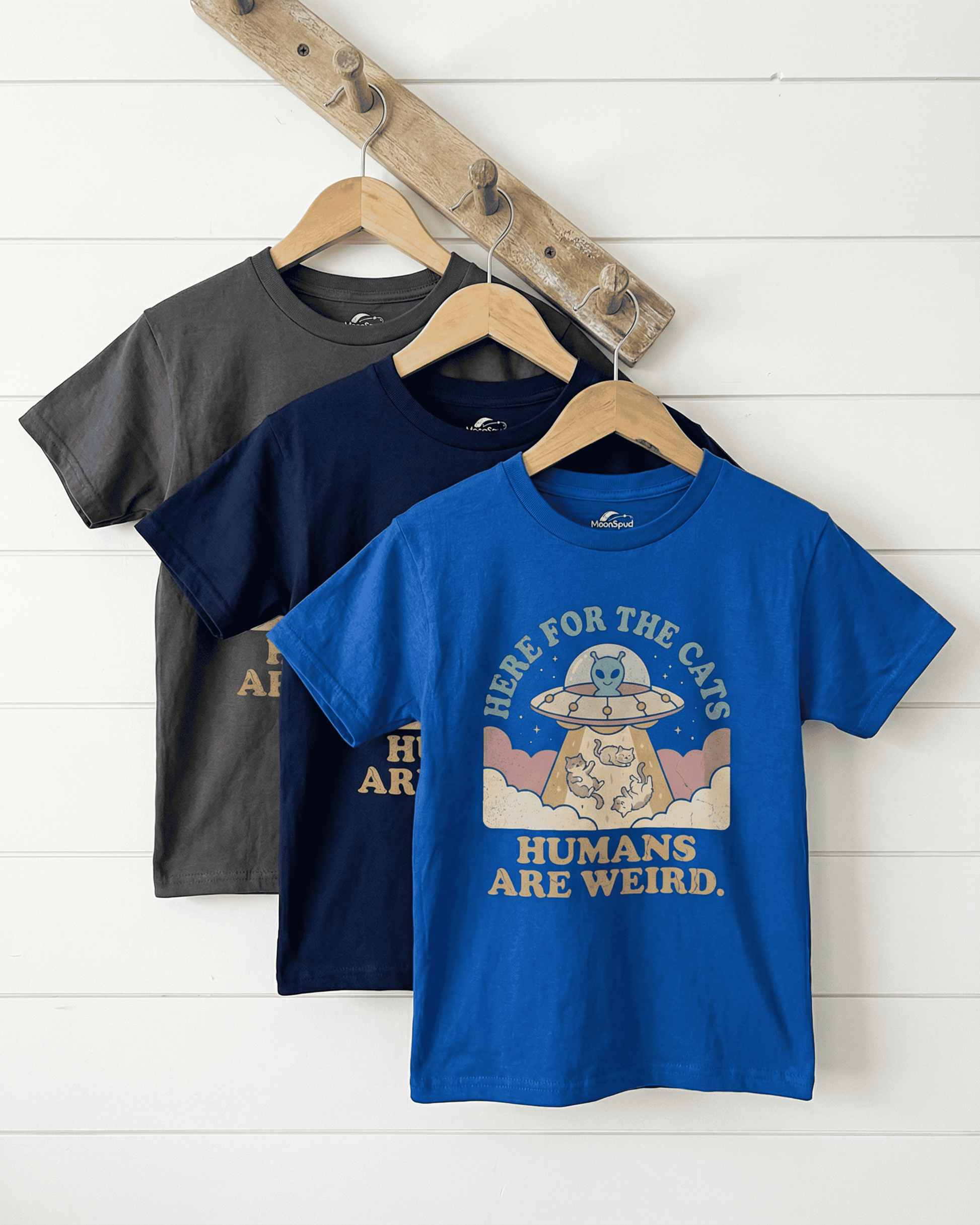 A royal blue kids 'UFO Here for the Cats' graphic tee hangs on a rustic wooden rack. This cute kids alien cat t-shirt design is shown up close, with the charcoal and navy versions visible in the background.